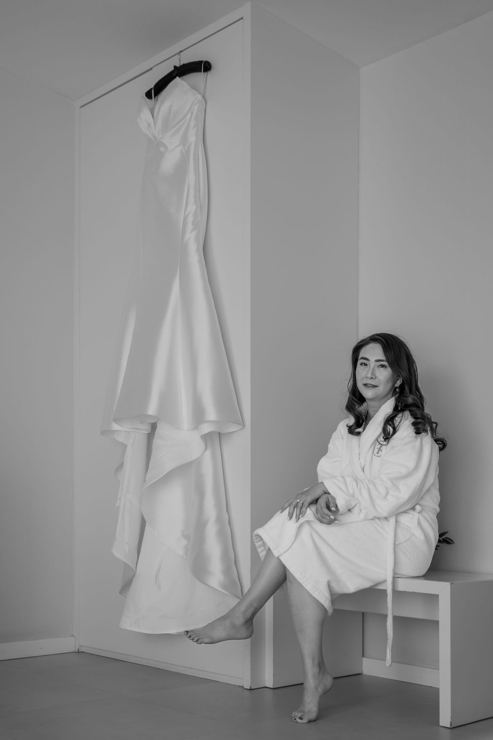 A wedding dress hangs on the wall while someone in a white robe sits on a white bench in a minimalist room.