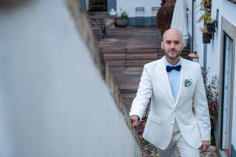 A groom in a white tuxedo jacket and blue bowtie walks along a path outside a building.