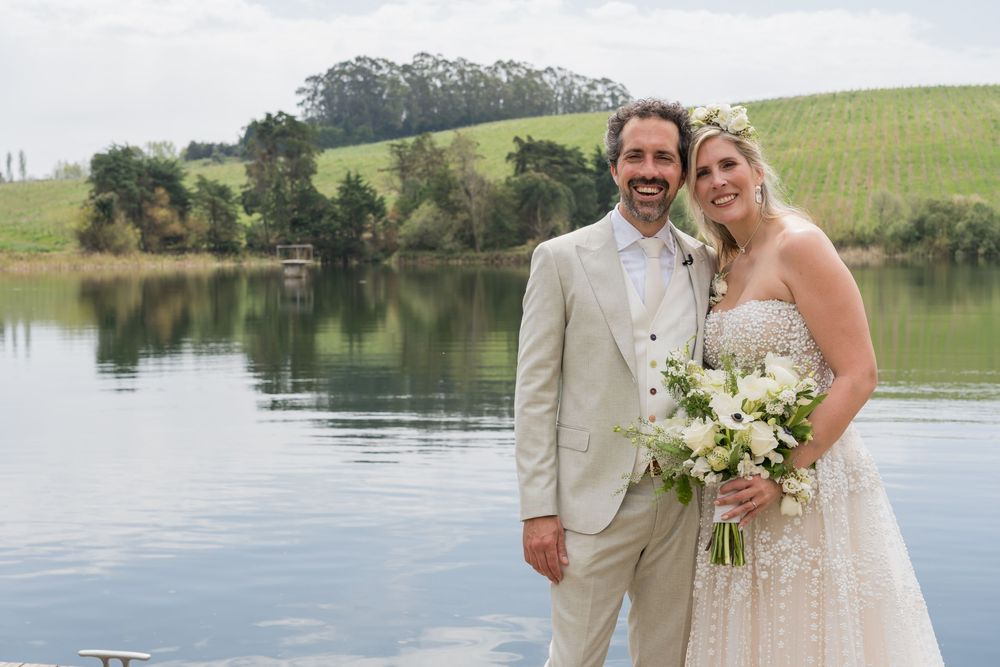 A couple in wedding attire poses by a serene lake with rolling green hills in the background.