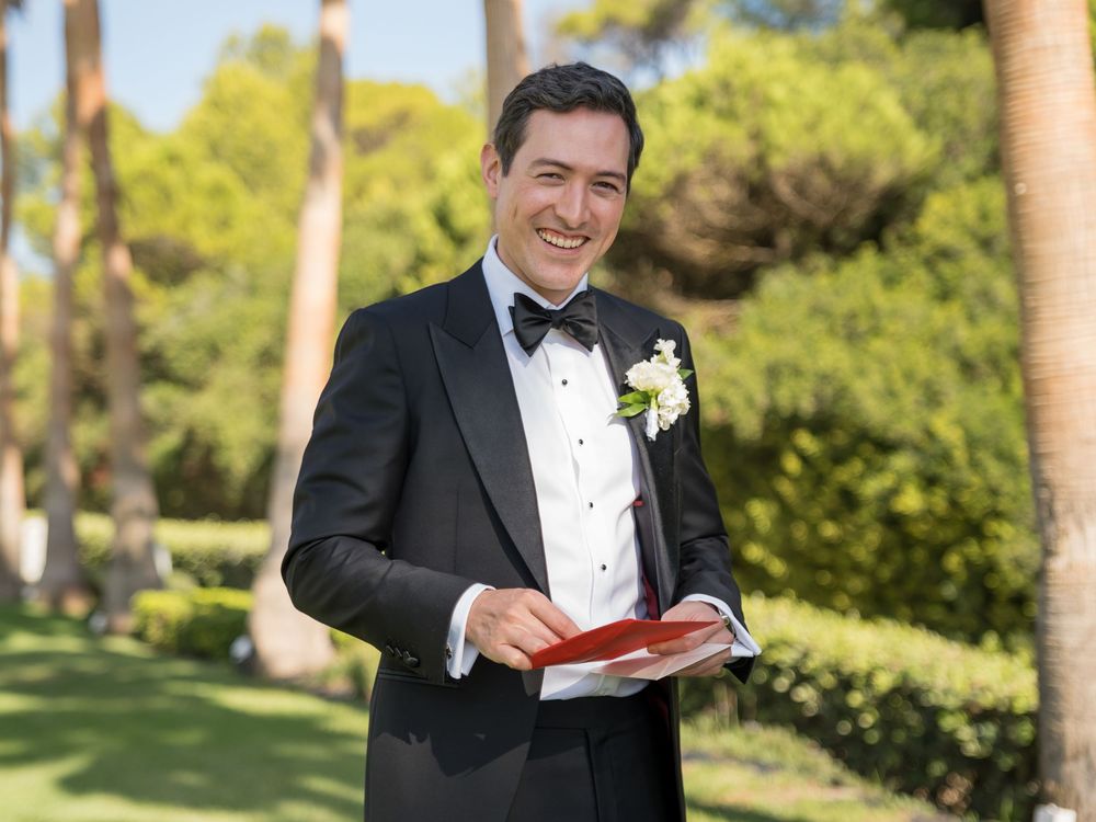 Someone in a black tuxedo smiles while reading a book outdoors in a sunny garden setting.