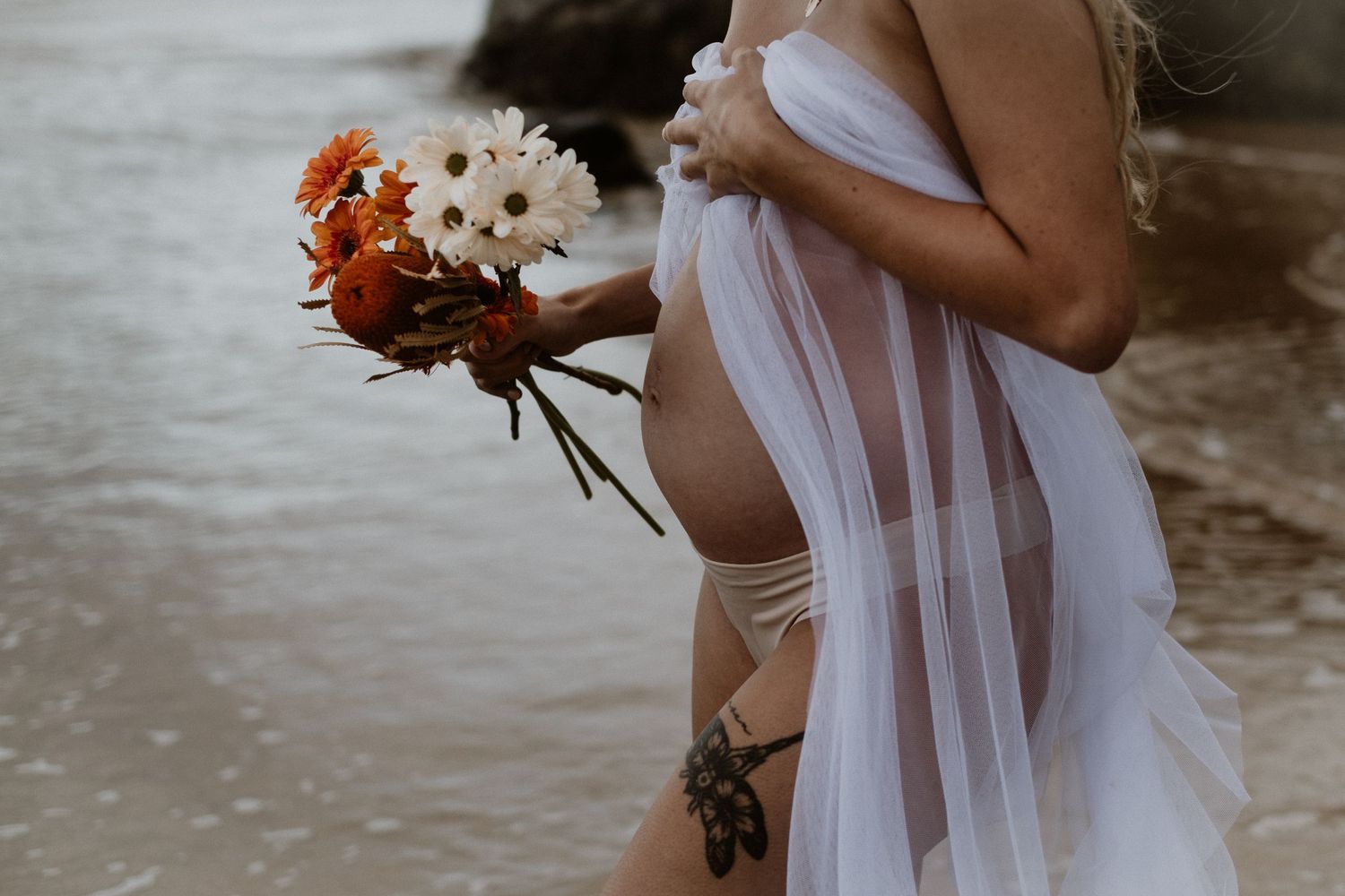 A person in flowing white fabric holds a bouquet of flowers on a rocky beach.