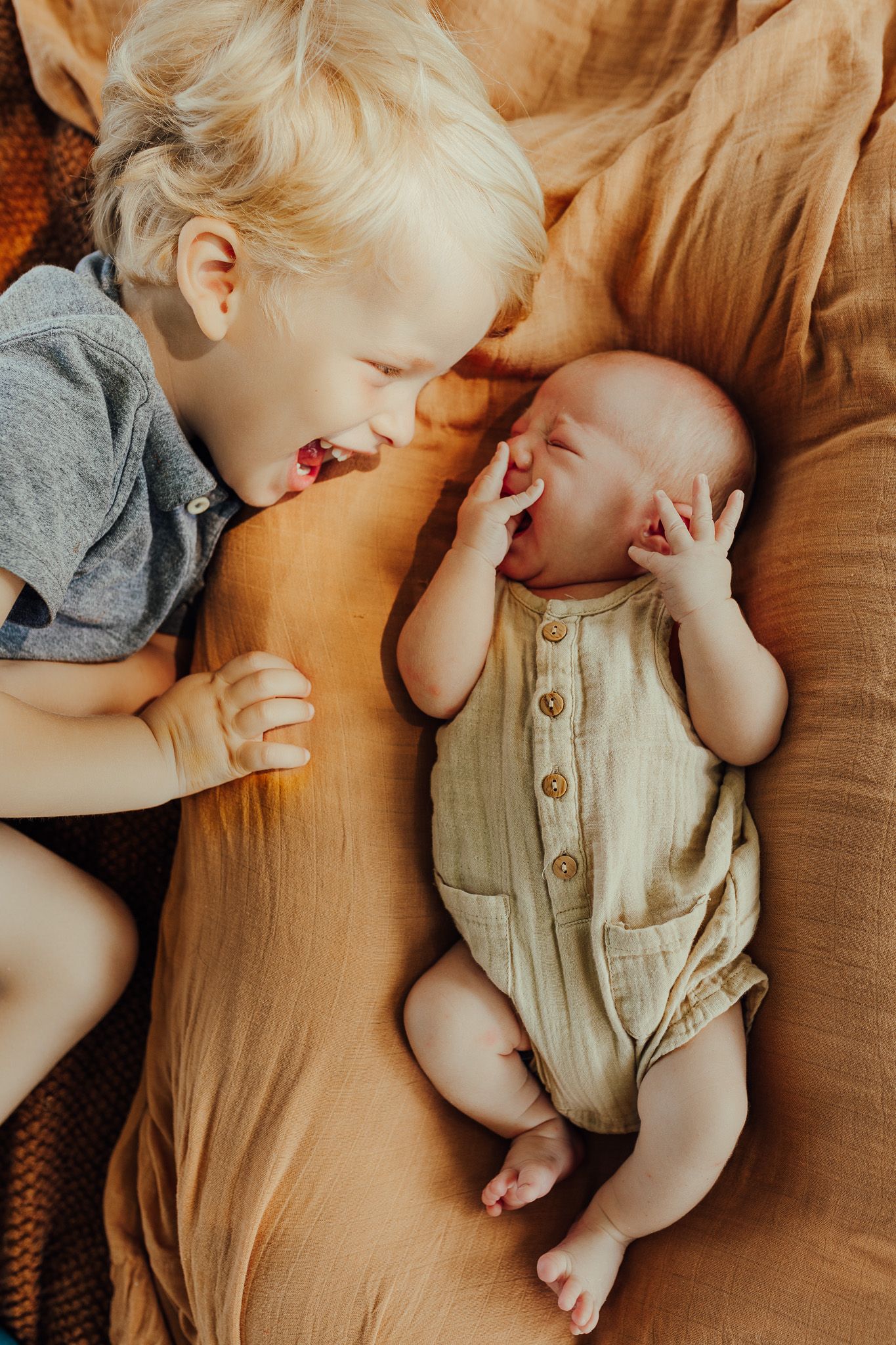 Two young children lying together on tan bedding, with one wearing grey and the other in a beige buttoned romper.
