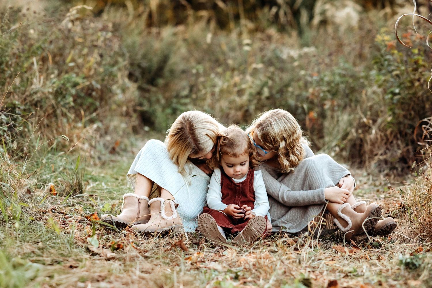 Three children in light clothing huddled together looking at a mobile phone while sitting in an autumn field.