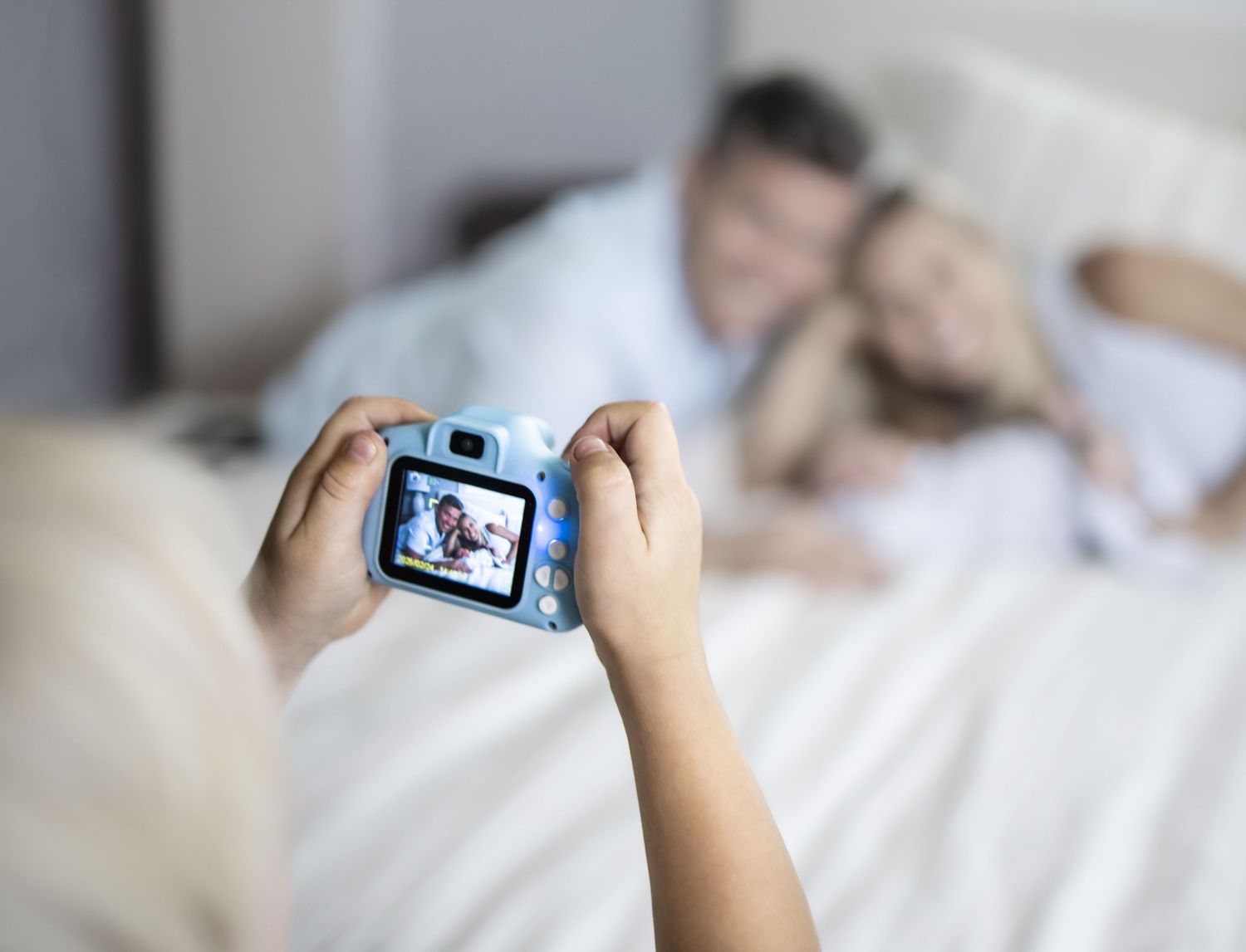 A little boy taking a photo with a toy digital camera of a couple resting on a bed with baby in soft focus.