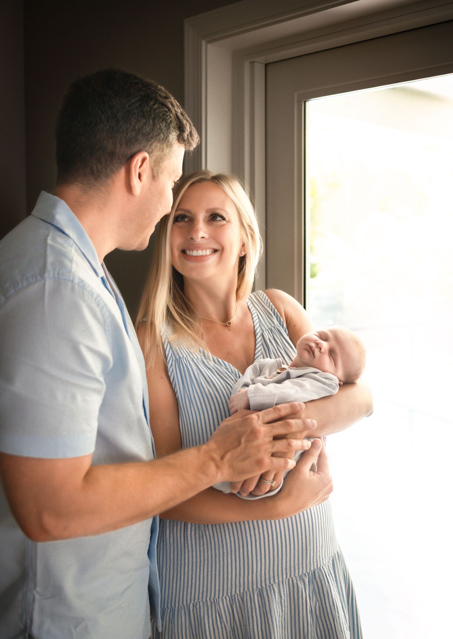 A couple in a bright room shares a tender moment while holding and gazing at their newborn baby. Lifestyle In-home Newborn Photography.
