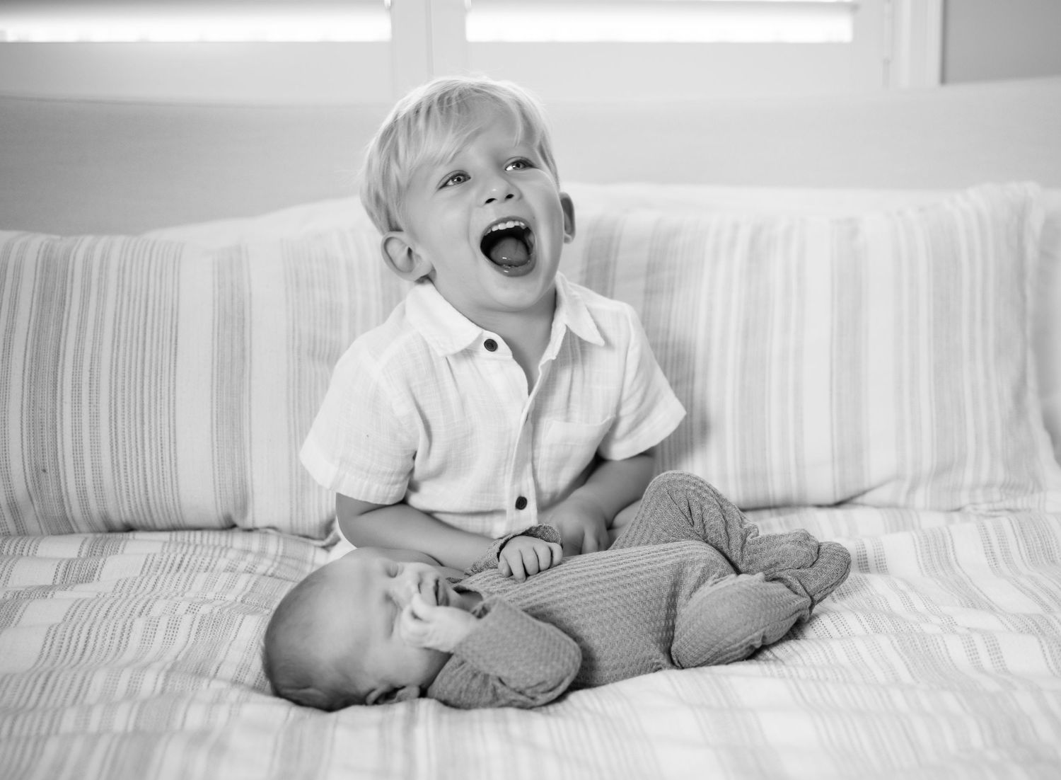 Black and white photo showing a toddler laughing next to newborn brother on a bed. Lifestyle In-home Newborn Photography.