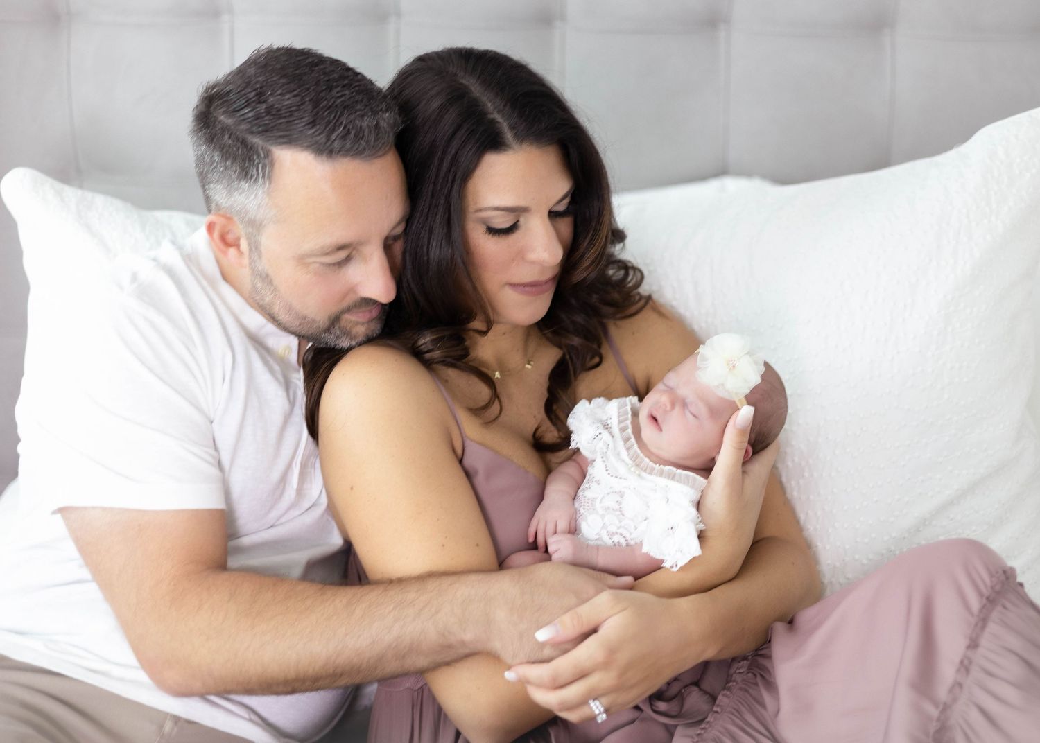 A couple lying in bed together holding and admiring their newborn baby wrapped in a white blanket.