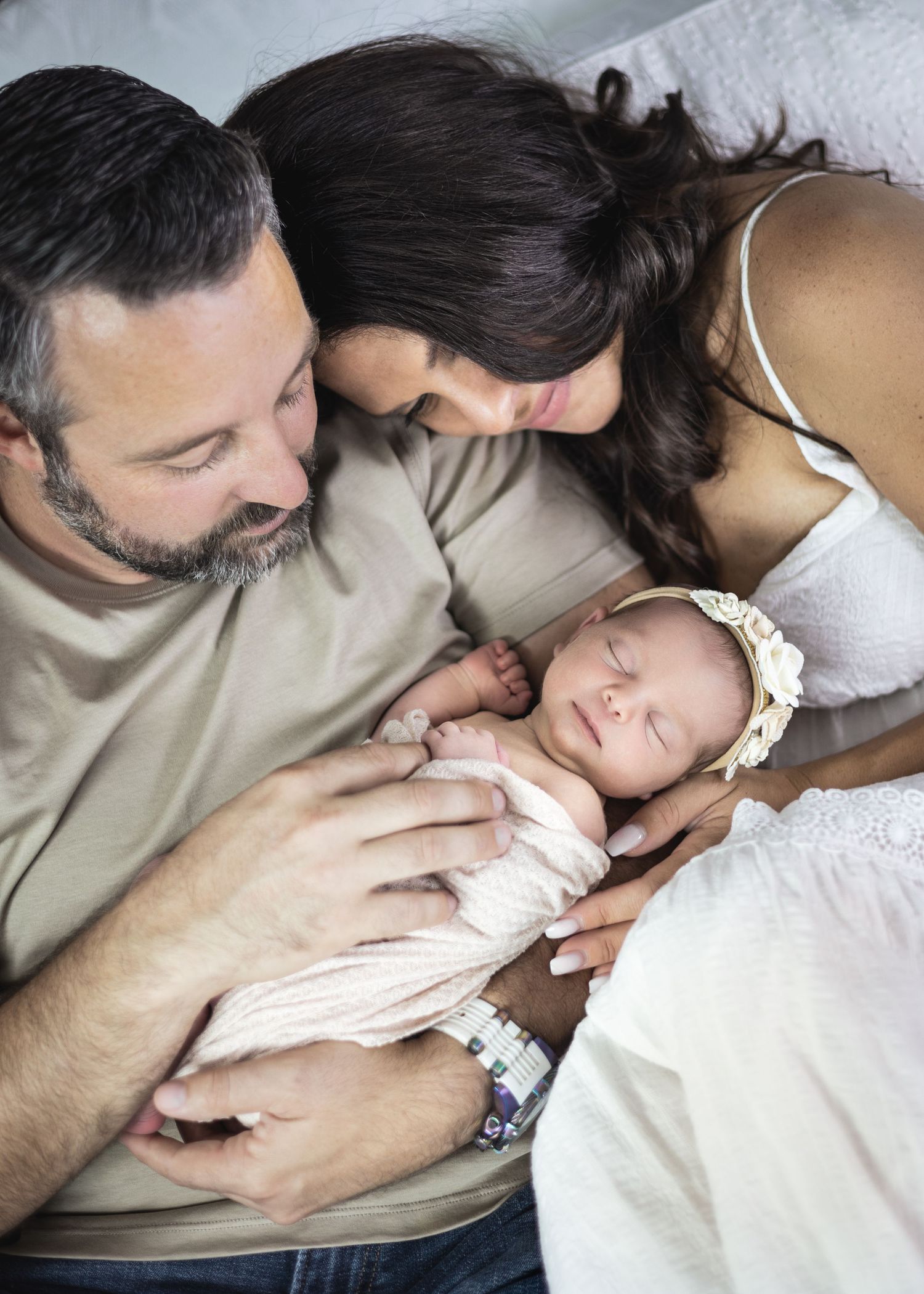 A sleeping newborn baby in white clothing is tenderly held by parents resting together on a bed.