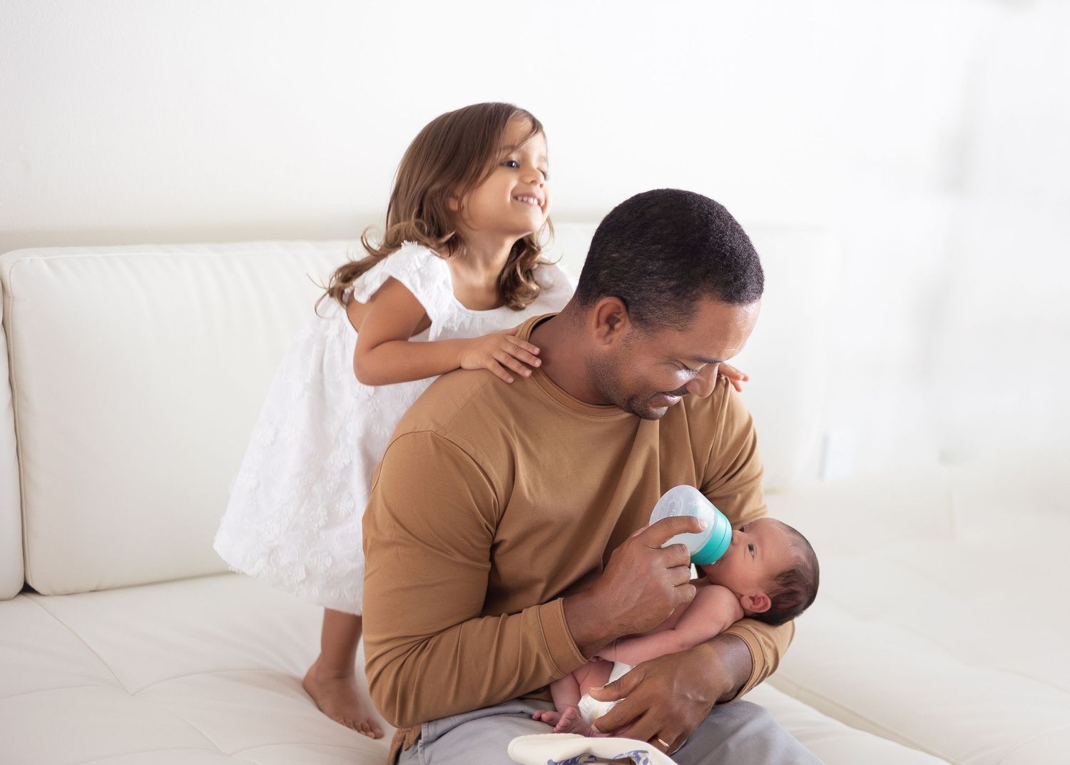 Dad, baby and big sister share tender moments on white couch in series of lifestyle photos.