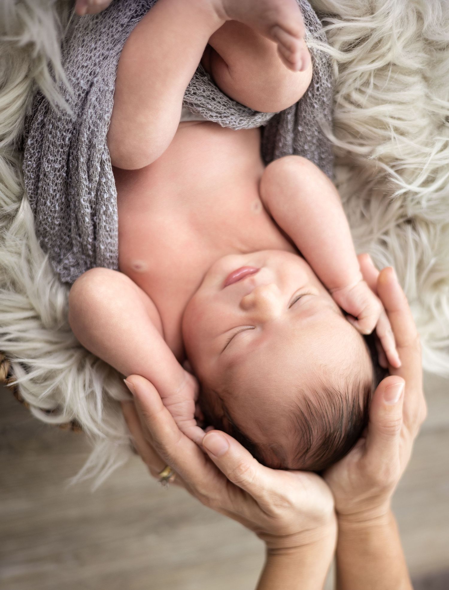 A newborn baby lying on a white fluffy rug while gentle hands cradle their head in a tender pose.