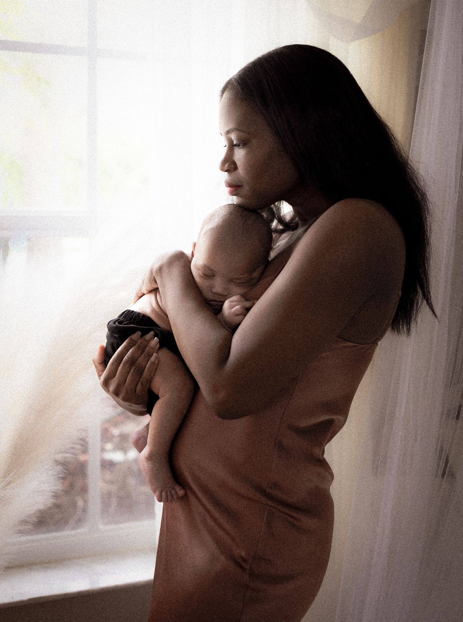 Mother in silhouette holds and comforts baby by window in emotional portrait series.