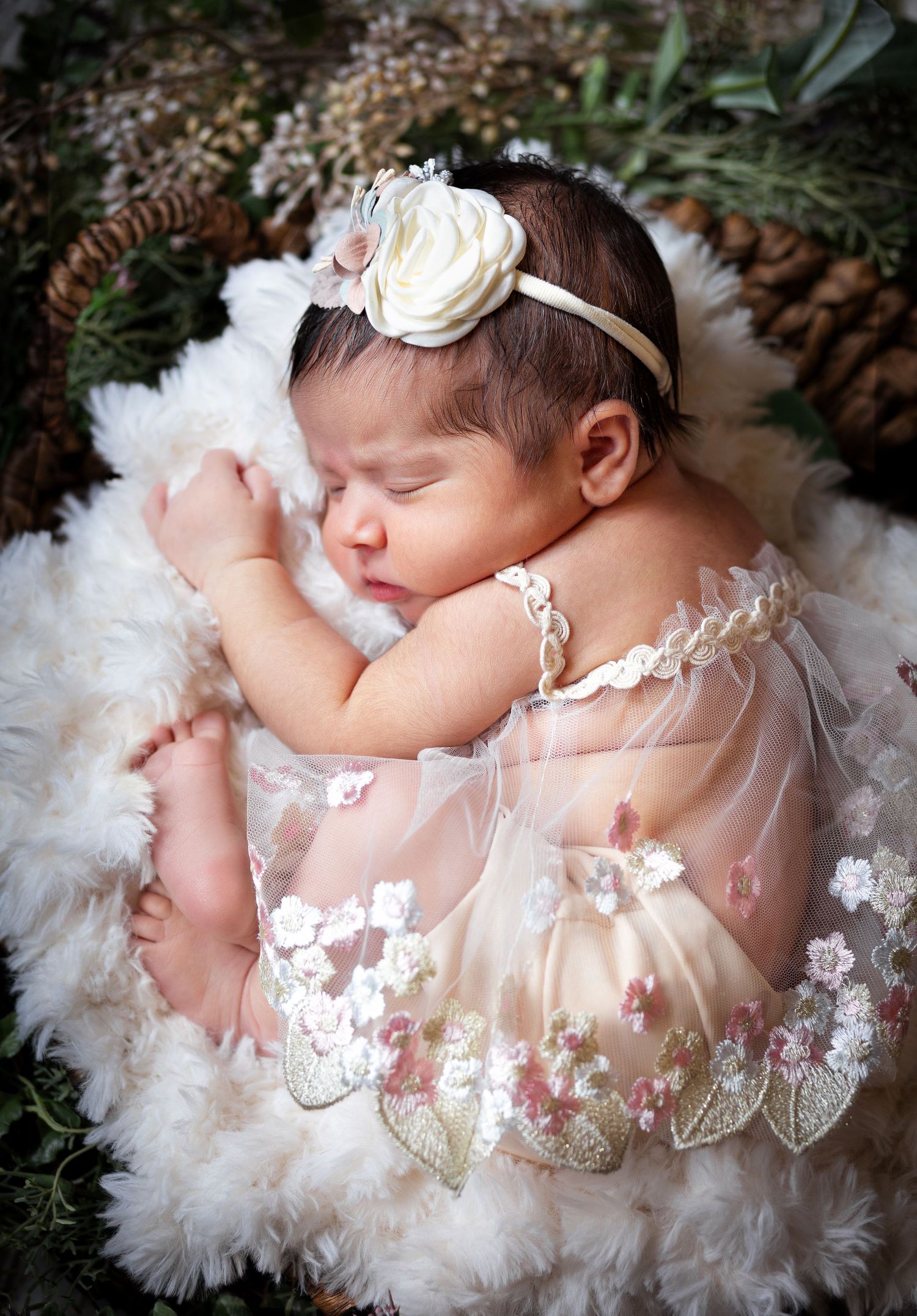 A sleeping newborn in a delicate white tulle dress and headband rests peacefully on a fluffy white blanket.