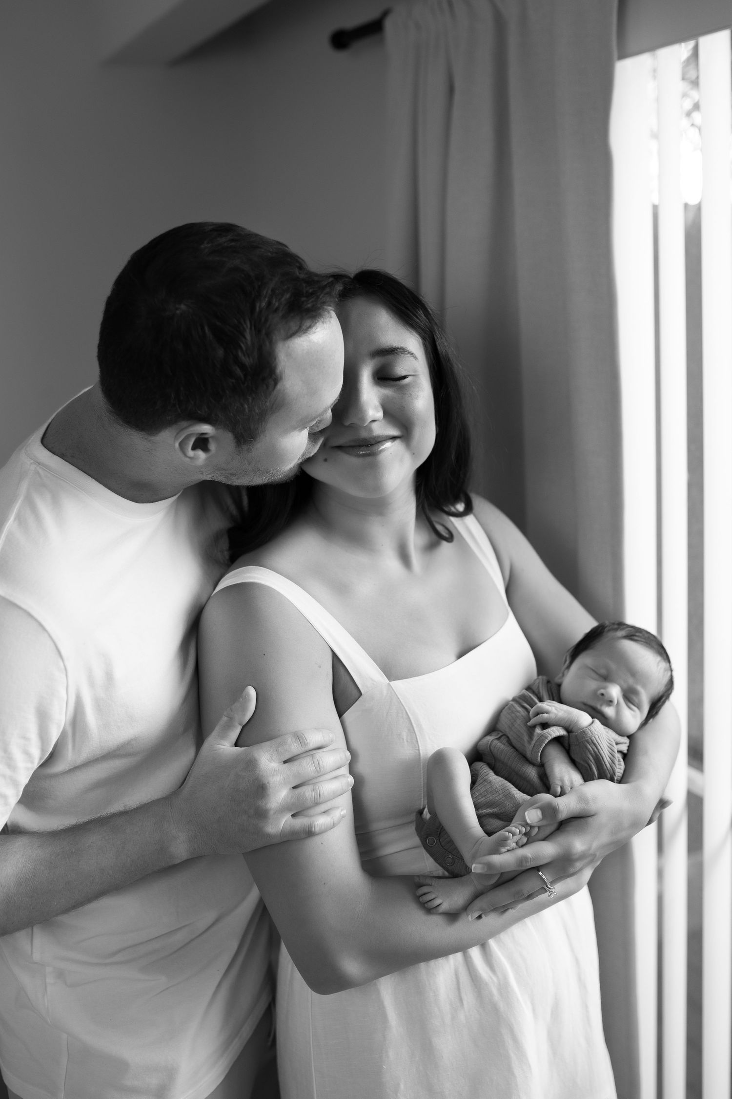 Black and white artistic photo showing intimate moments between a couple and newborn baby in white clothing by a window.