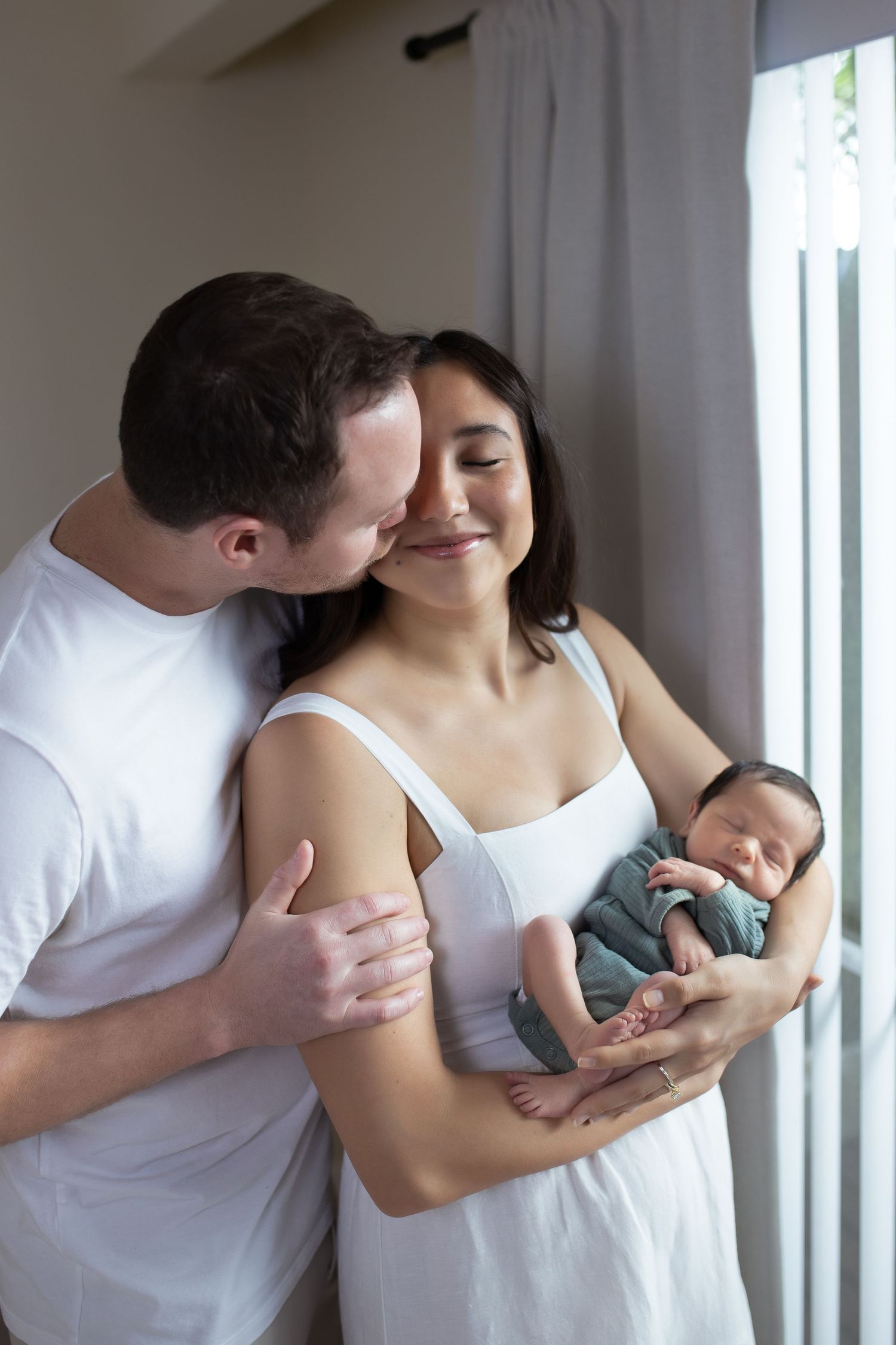 A series of tender intimate moments between a couple holding their newborn baby near a window.