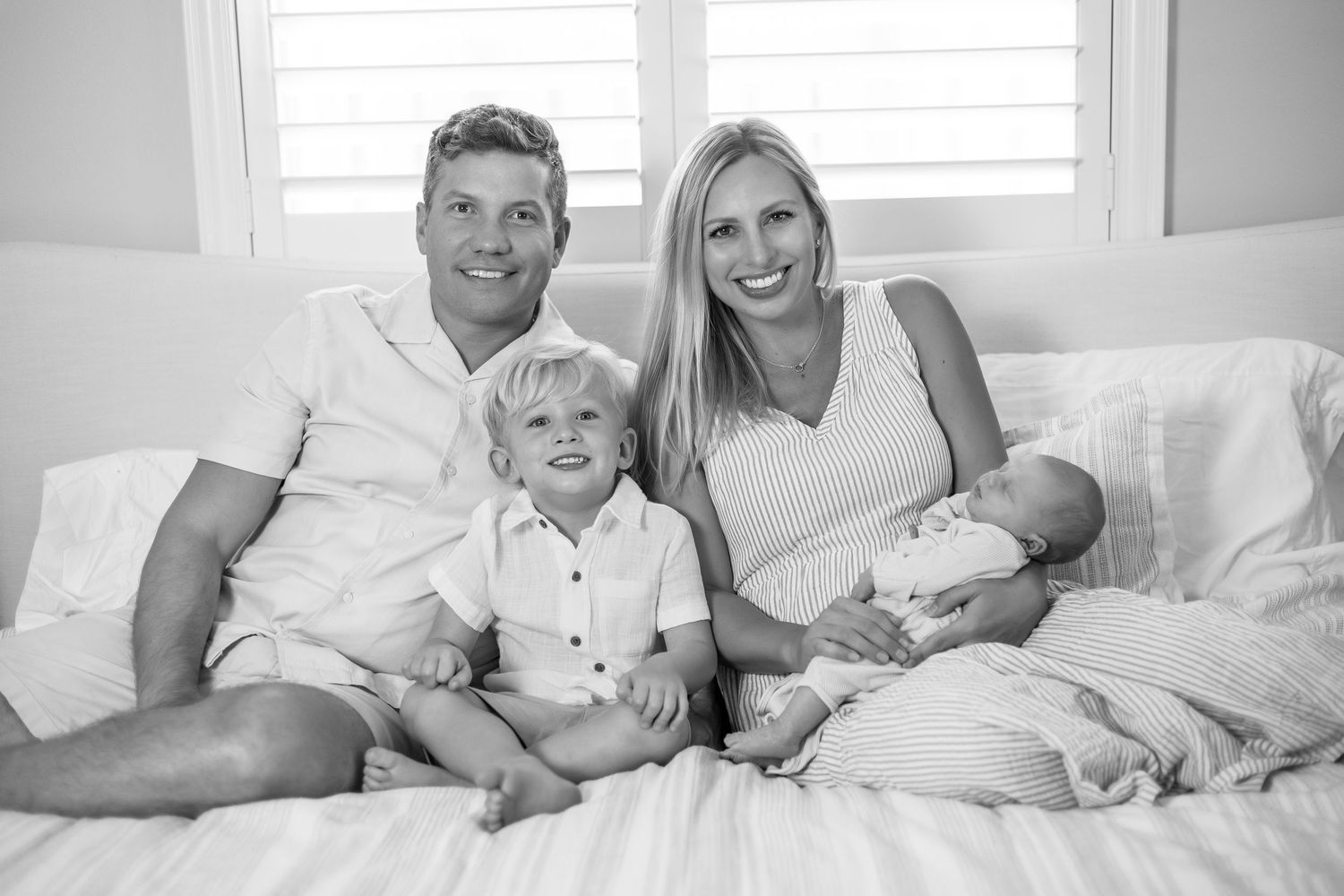 A black and white photo showing a family relaxing together on a white bed with newborn baby. Lifestyle In-home Newborn Photography.