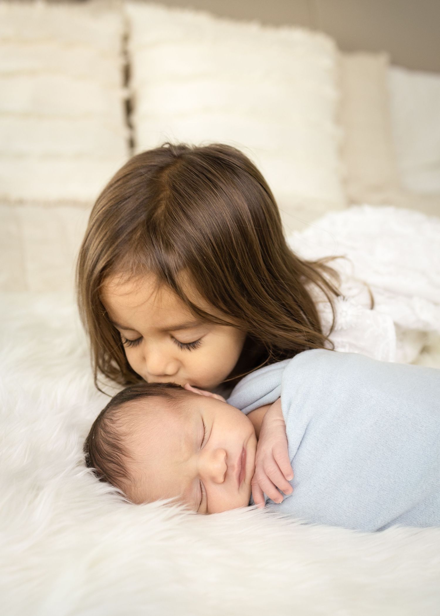 A tender moment as an older sibling gently kisses a sleeping newborn baby swaddled in blue on white bedding.