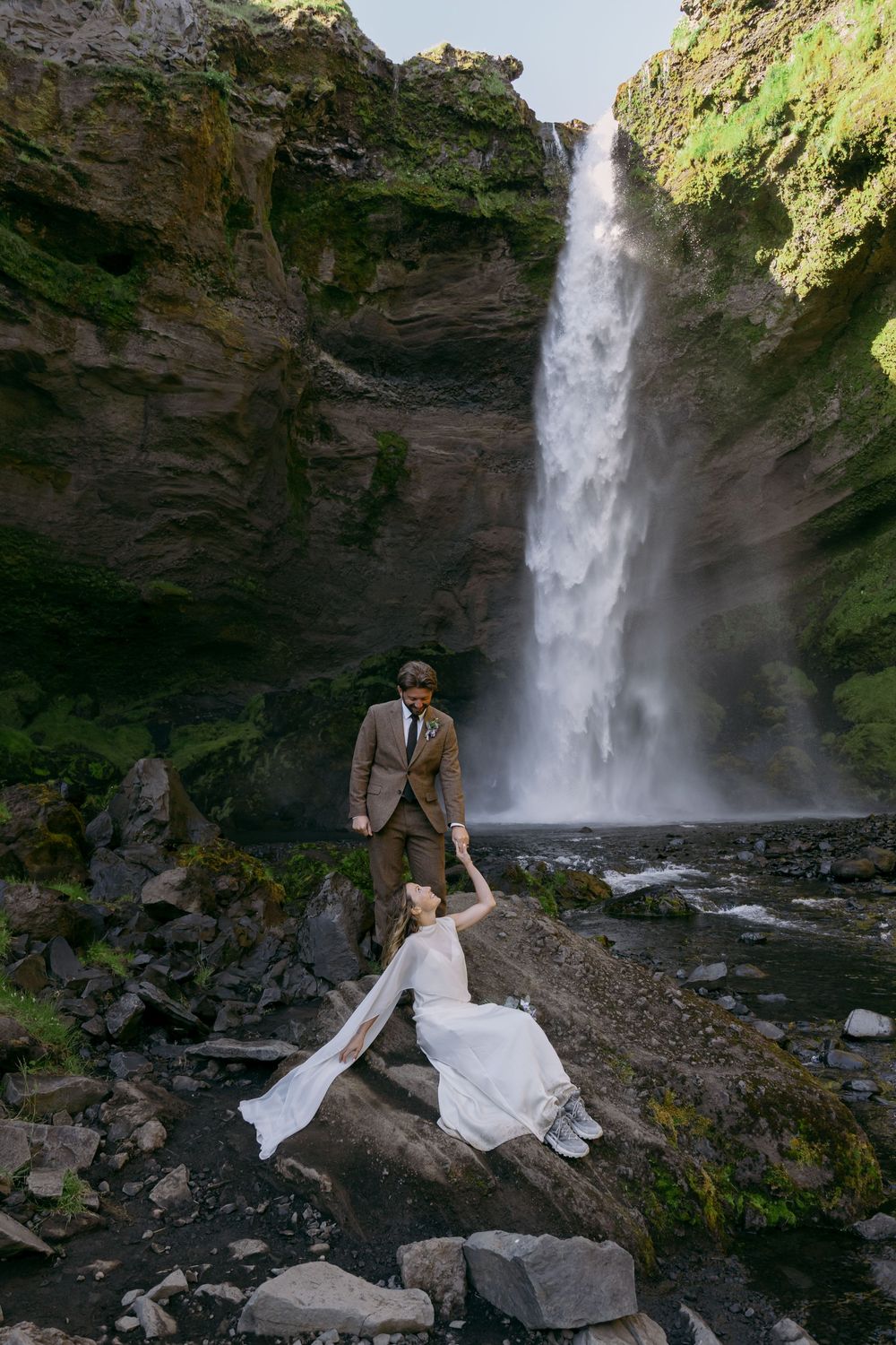 Bride and groom pose before a stunning Iceland waterfall, she sits on rocks in white gown as he stands holding her hand.
