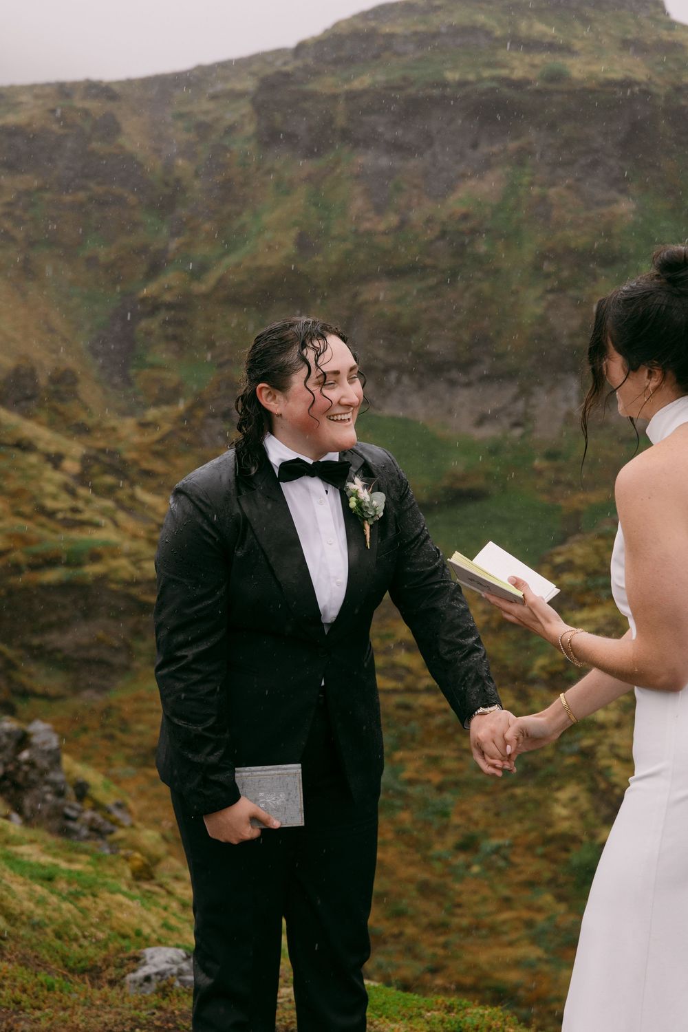 Couple exchanging vows outdoors in the rain on a misty green mountainside, groom in black tuxedo smiling joyfully.