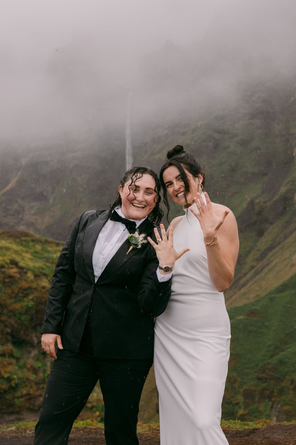 Two brides show off wedding rings outdoors near a misty waterfall, one in a tuxedo and one in a white gown.