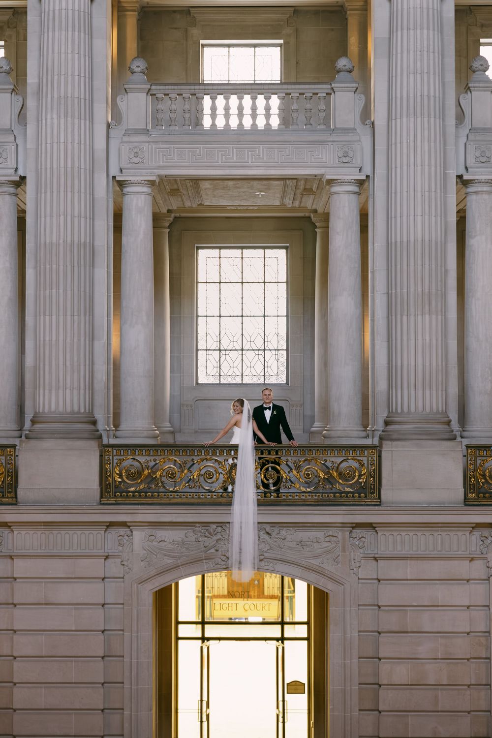 Bride and groom stand on ornate balcony of San Francisco City Hall, framed by grand columns and arched doorway below.