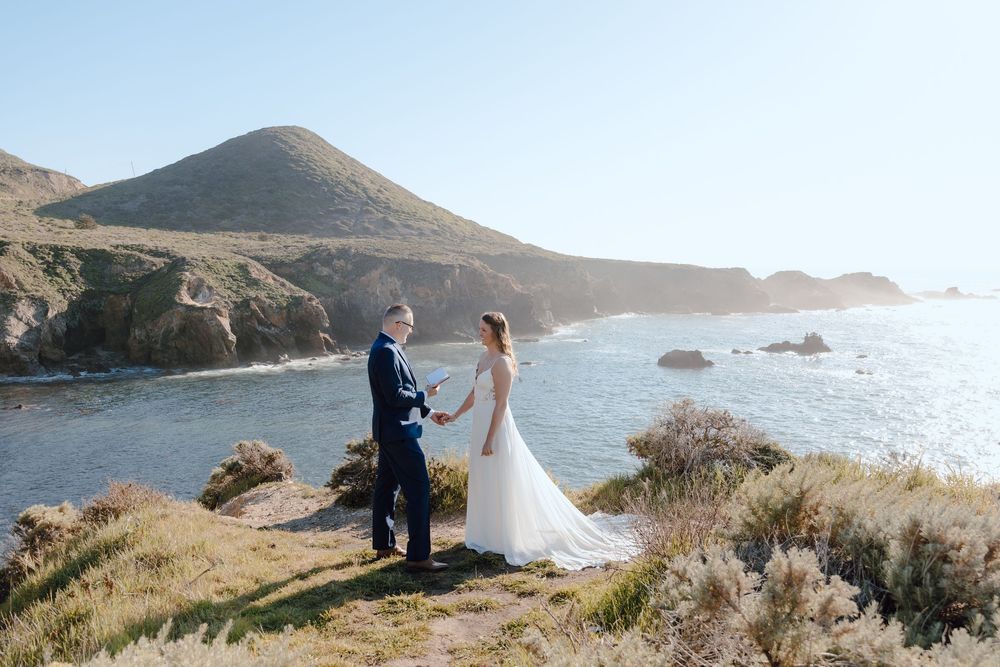 Couple exchanging vows at a stunning coastal cliffside wedding with ocean views and rolling hills in the background.