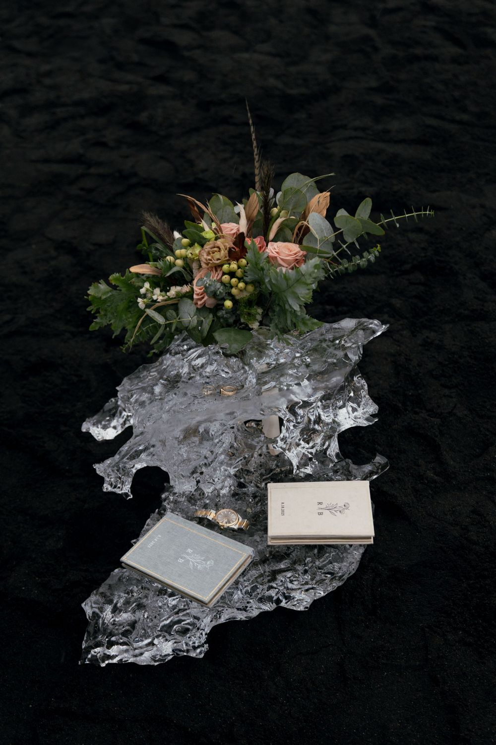 Floral bouquet with roses and greenery on ice shard with books and rings on dark rocky surface.