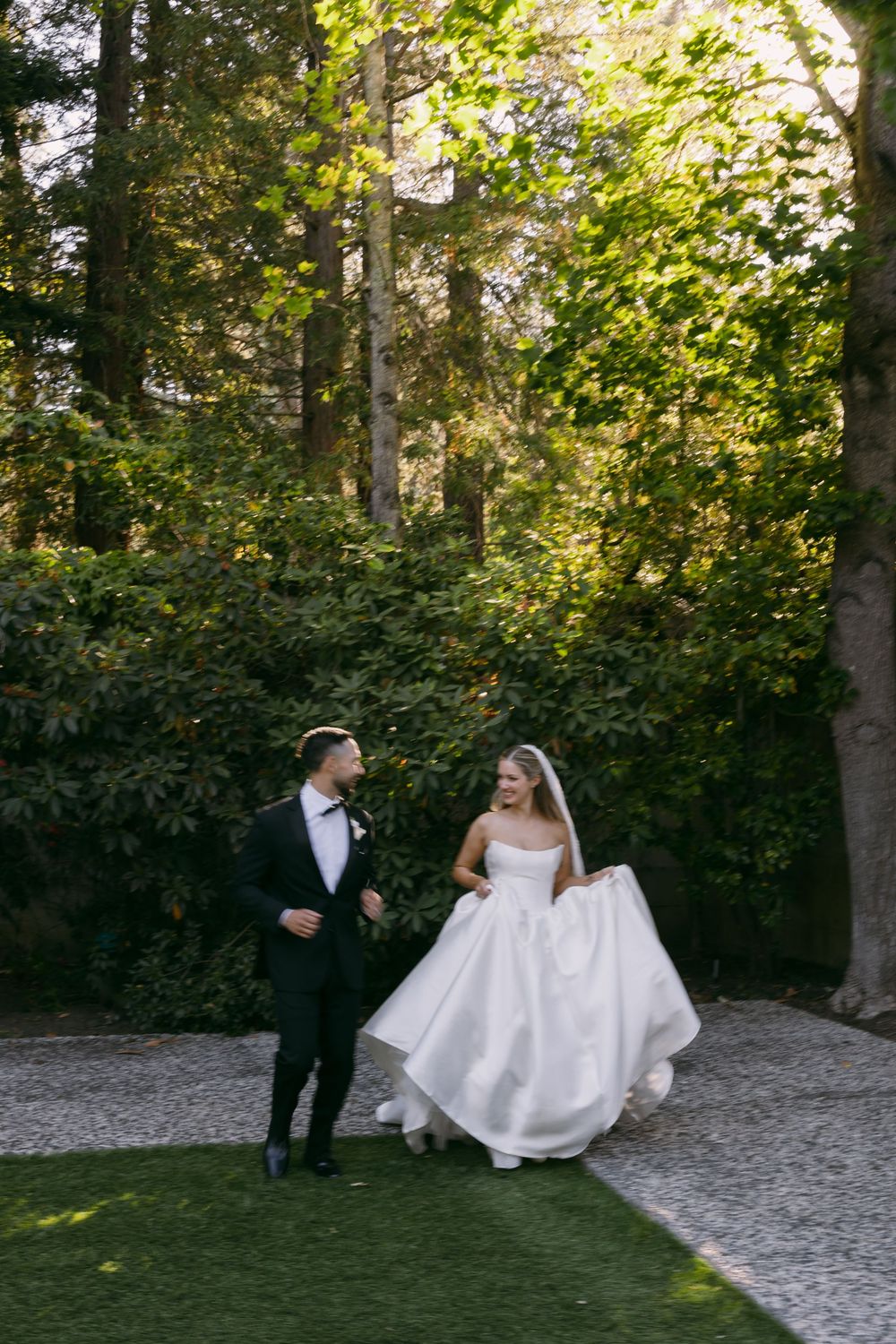 Bride in white ball gown and groom in black tuxedo walk hand in hand through lush green garden path.