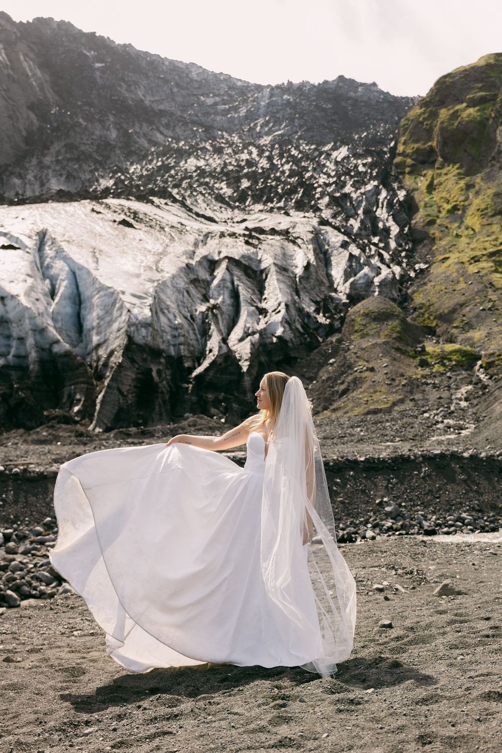 Bride in white gown and veil poses before a dramatic glacier and black sand landscape in Iceland.