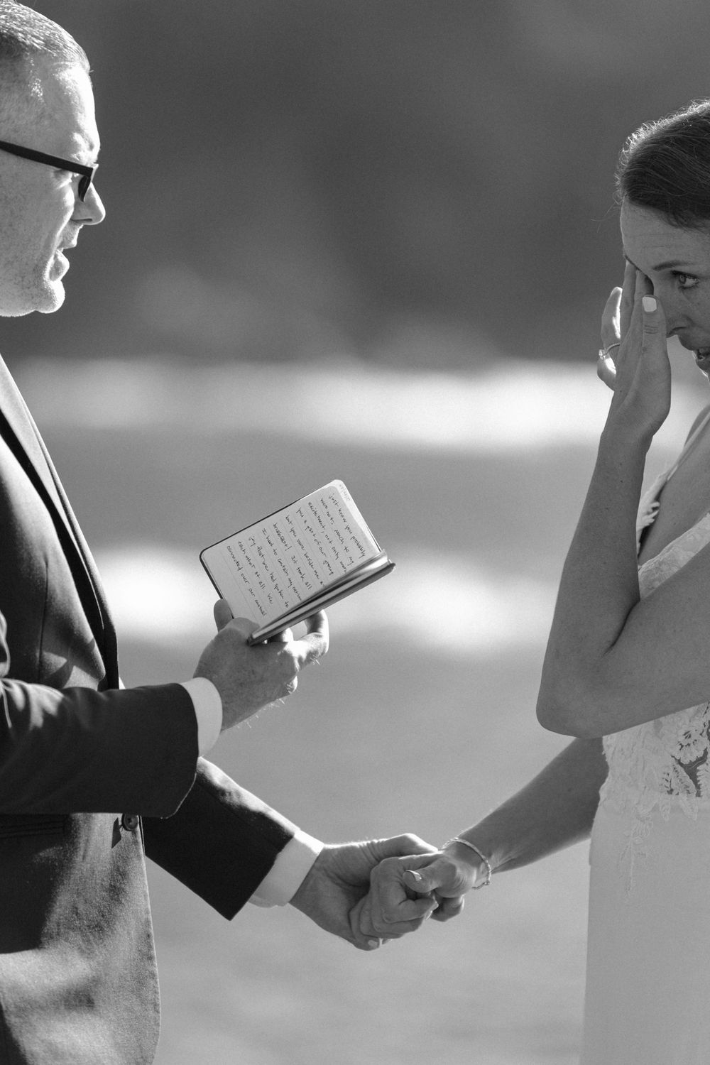 Black and white photo of groom reading vows from book while emotional bride wipes tears.