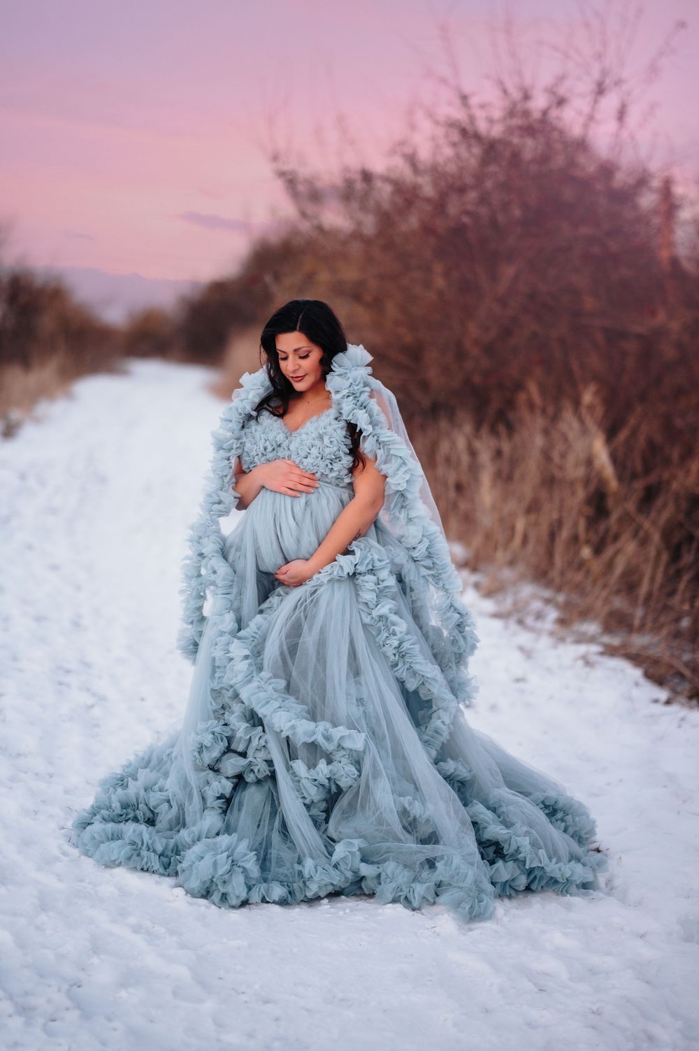 Ethereal maternity photo in a flowing blue ruffled gown against a snowy winter landscape at sunset.