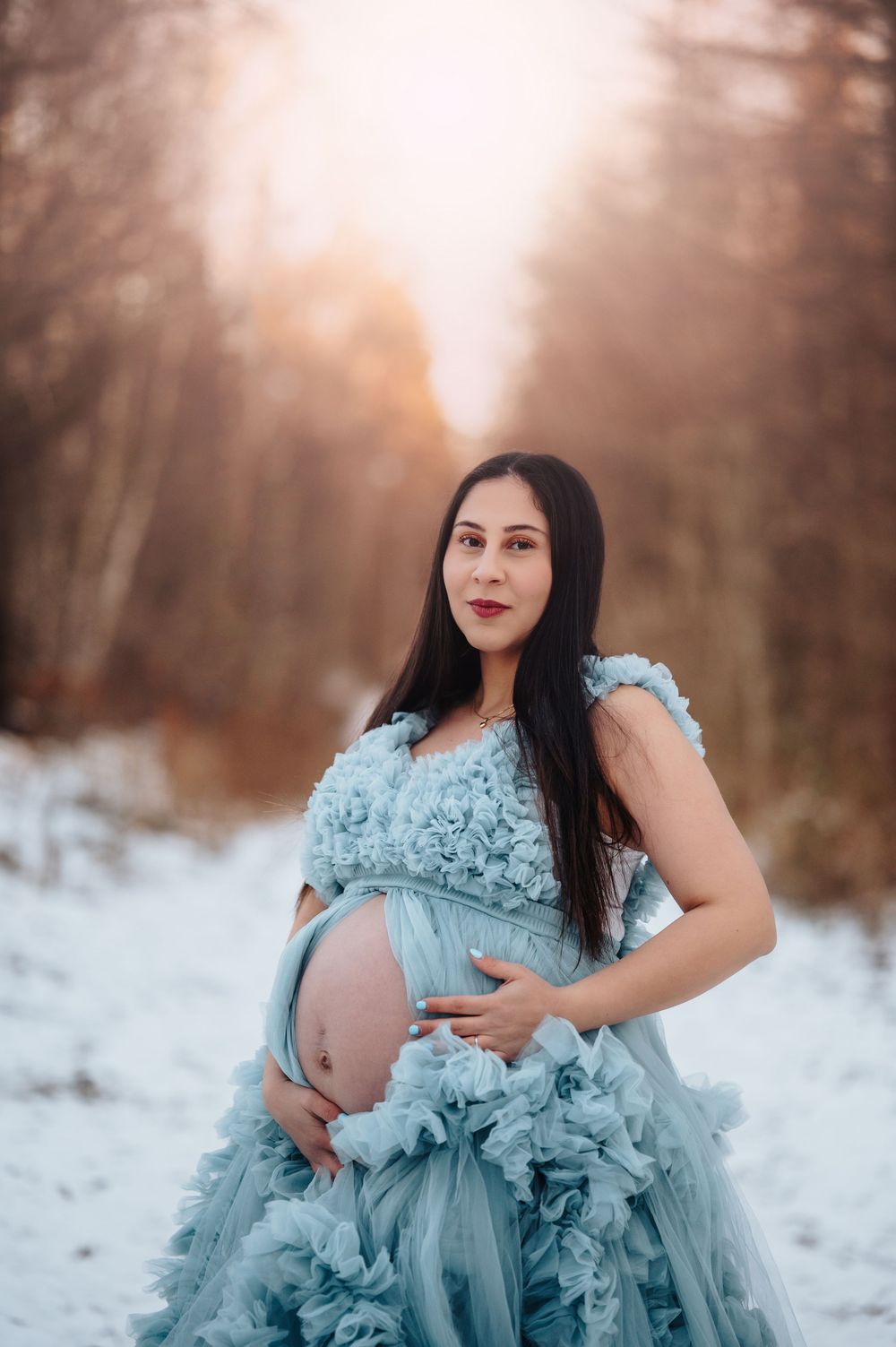 Maternity portrait in pale blue gown against snow-covered winter forest backdrop.