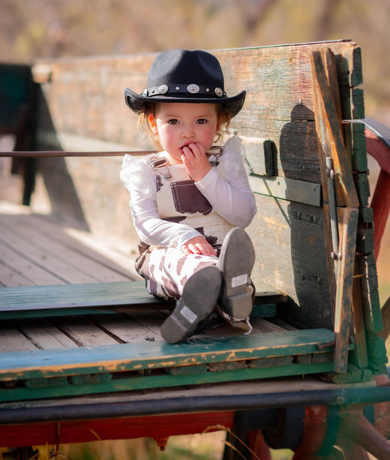 Adorable toddler in a black cowboy hat and overalls sitting on a rustic wooden wagon outdoors.