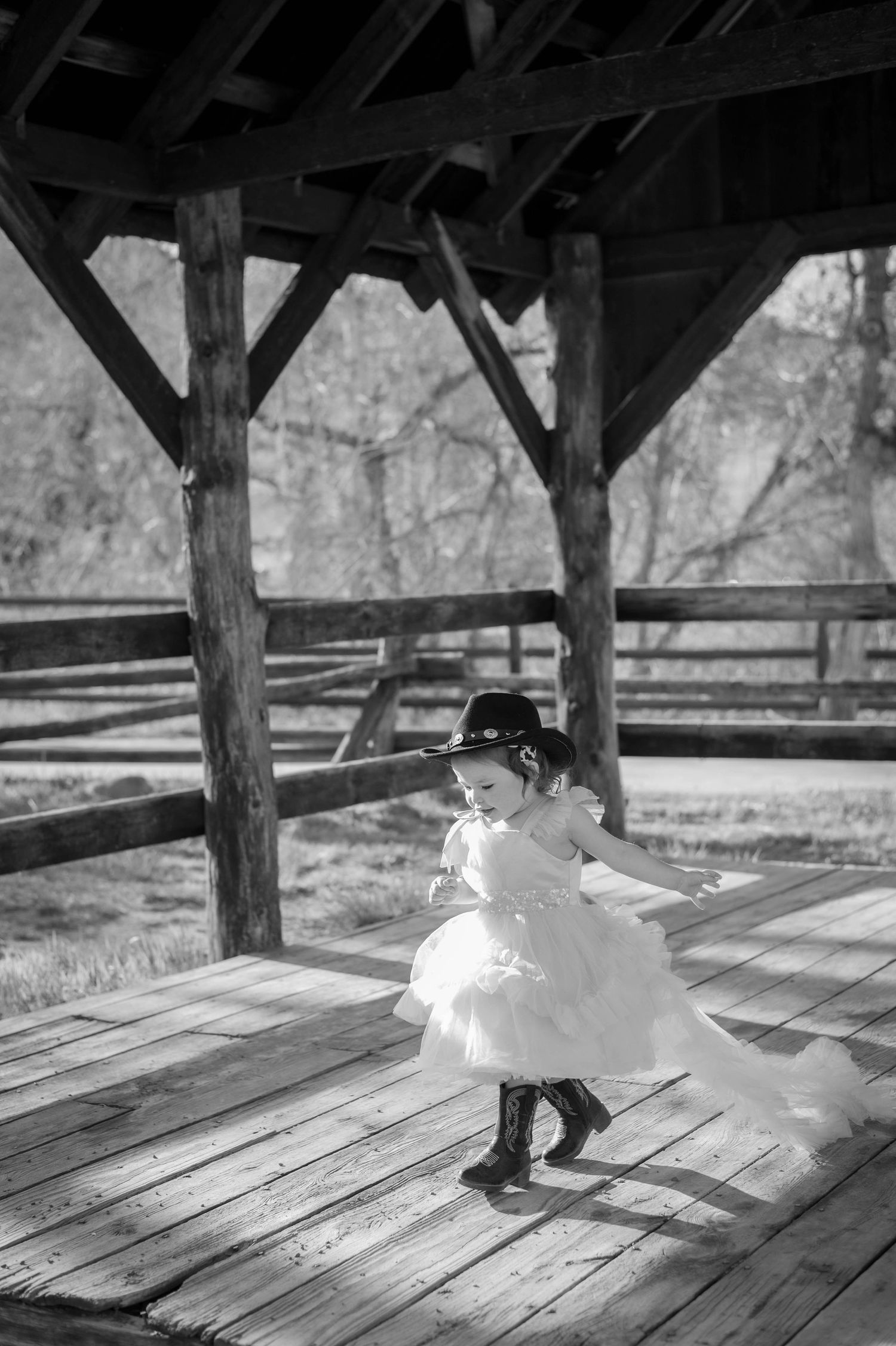 A little girl in a white tutu dress and cowboy hat dances on a wooden deck in a black and white photo.