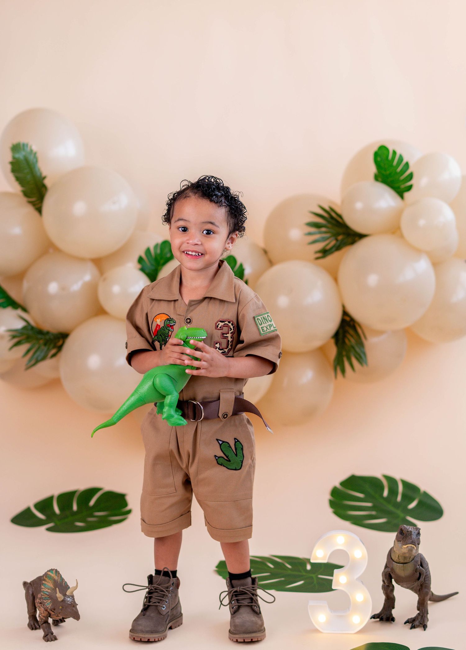 Toddler in safari outfit holds green dinosaur toy at dinosaur-themed 3rd birthday party with balloon arch.