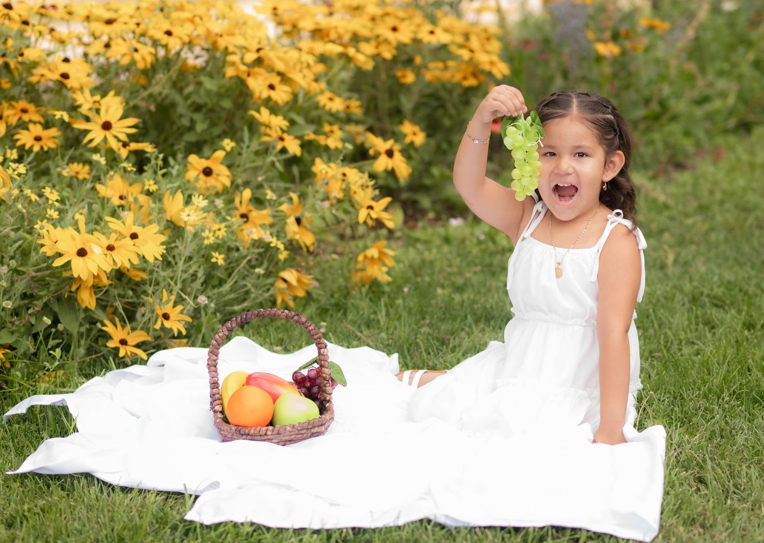 A cheerful little girl in a white dress sits on a blanket holding green grapes near a fruit basket with yellow flowers behind her.