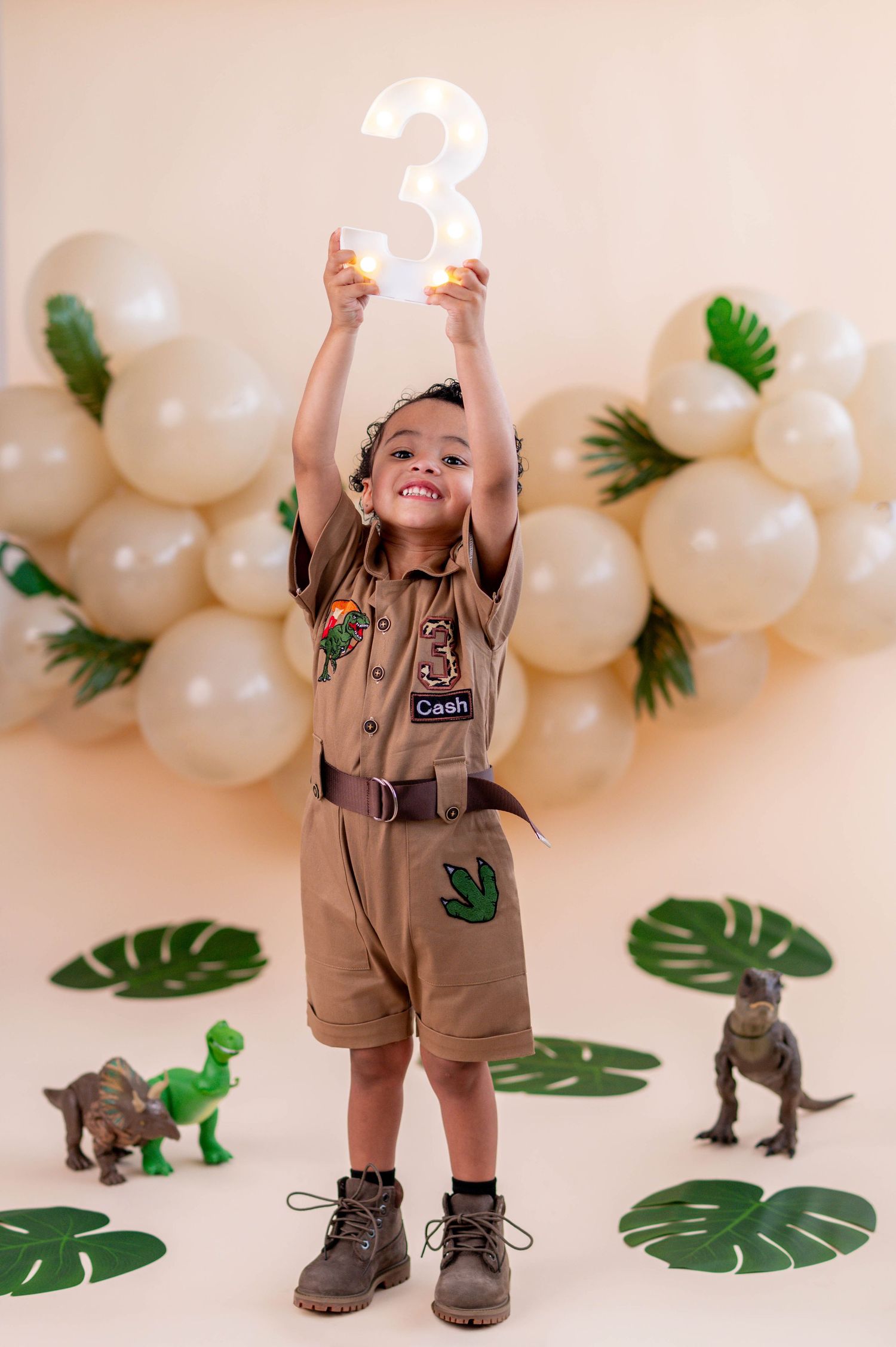A smiling boy in safari outfit holds a glowing number 3, surrounded by dinosaur toys and balloons at his birthday party.