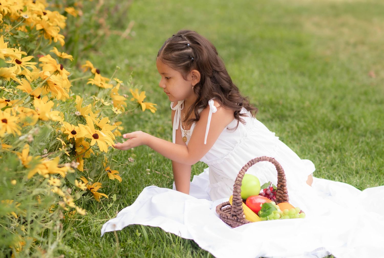 Young girl in white dress sitting on blanket in green field, reaching to touch yellow wildflowers beside a fruit basket.