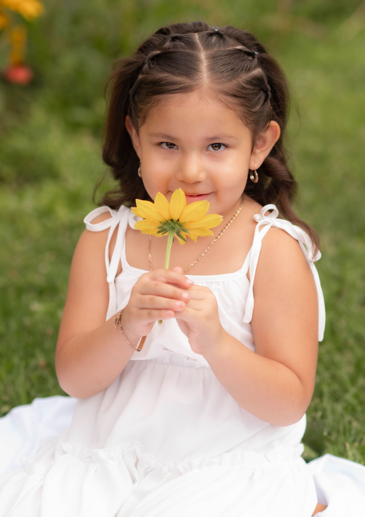 Young girl in white dress holding a yellow sunflower outdoors, smiling sweetly in a lush green garden setting.