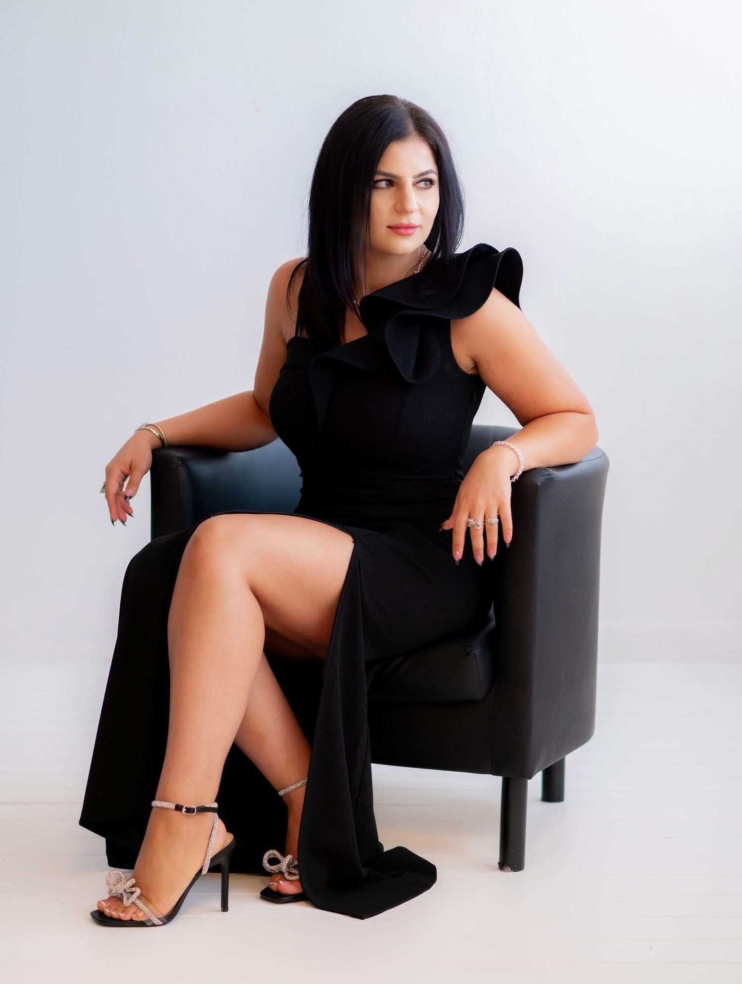 Elegant portrait session featuring black dress and chair against minimalist white backdrop.