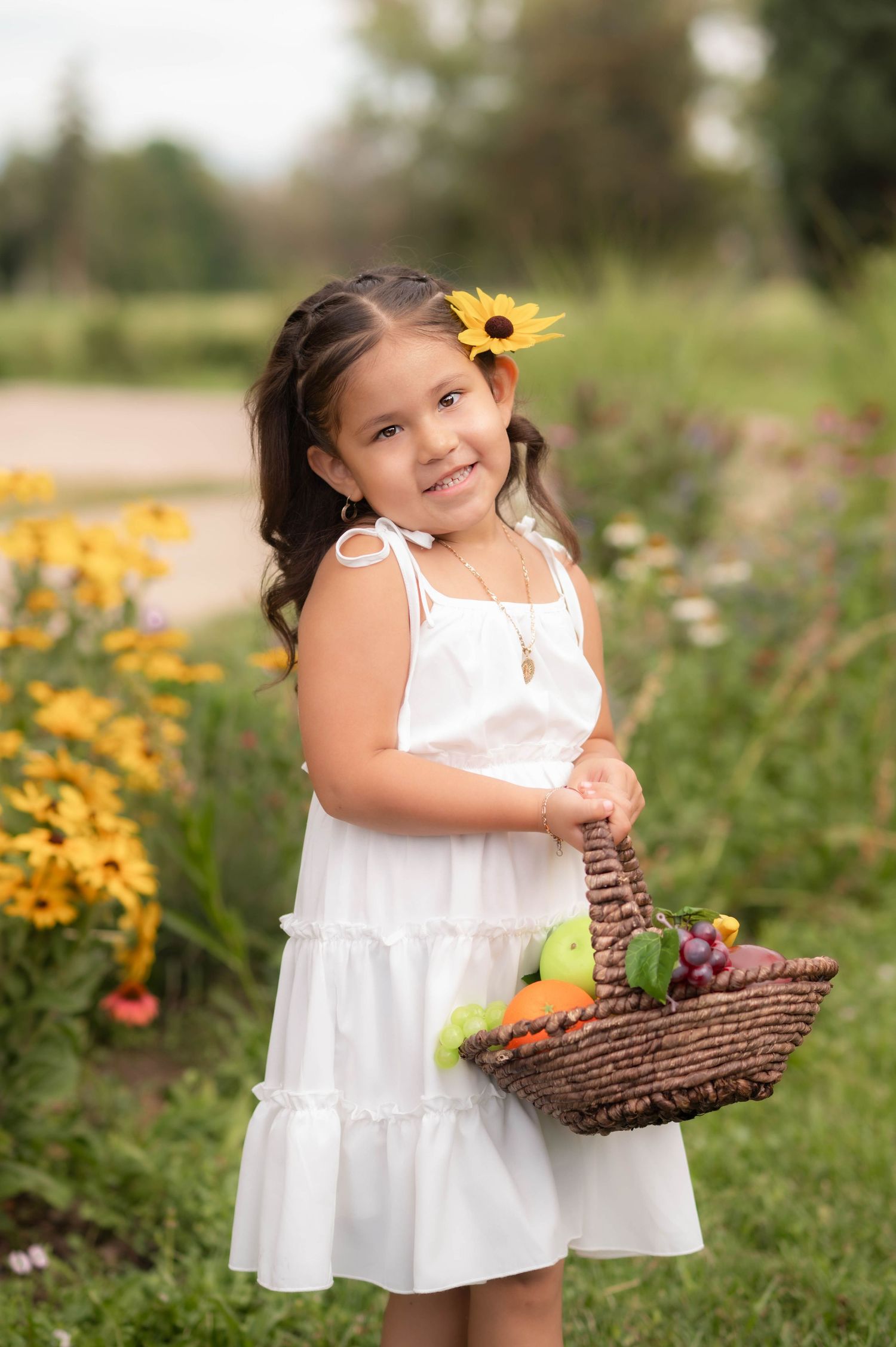 Young girl in white dress holding fruit basket with sunflower hair accessory, smiling in a colorful garden.