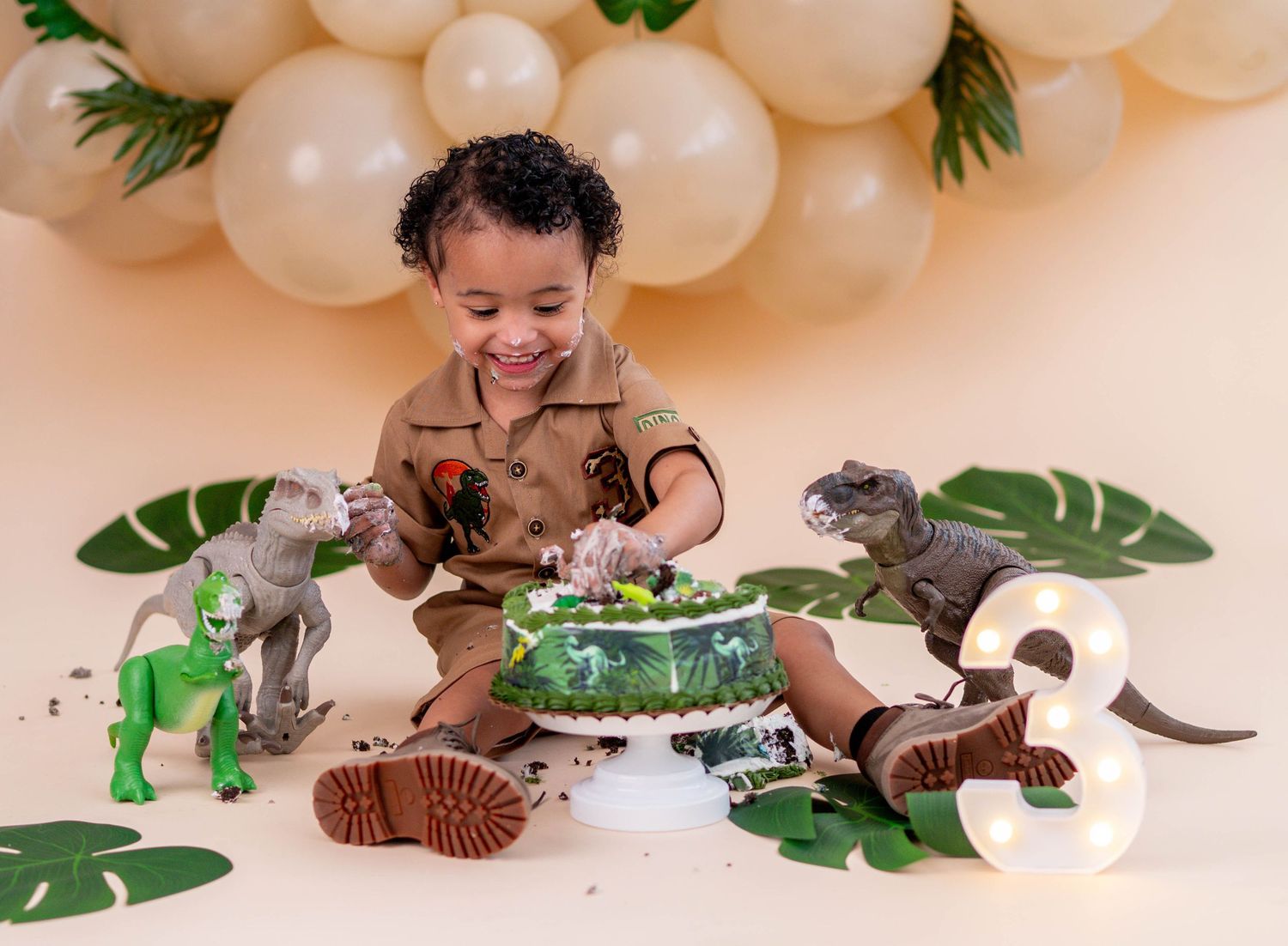 Toddler boy celebrating 3rd birthday with dinosaur themed cake smash, surrounded by toy dinosaurs and balloon arch.