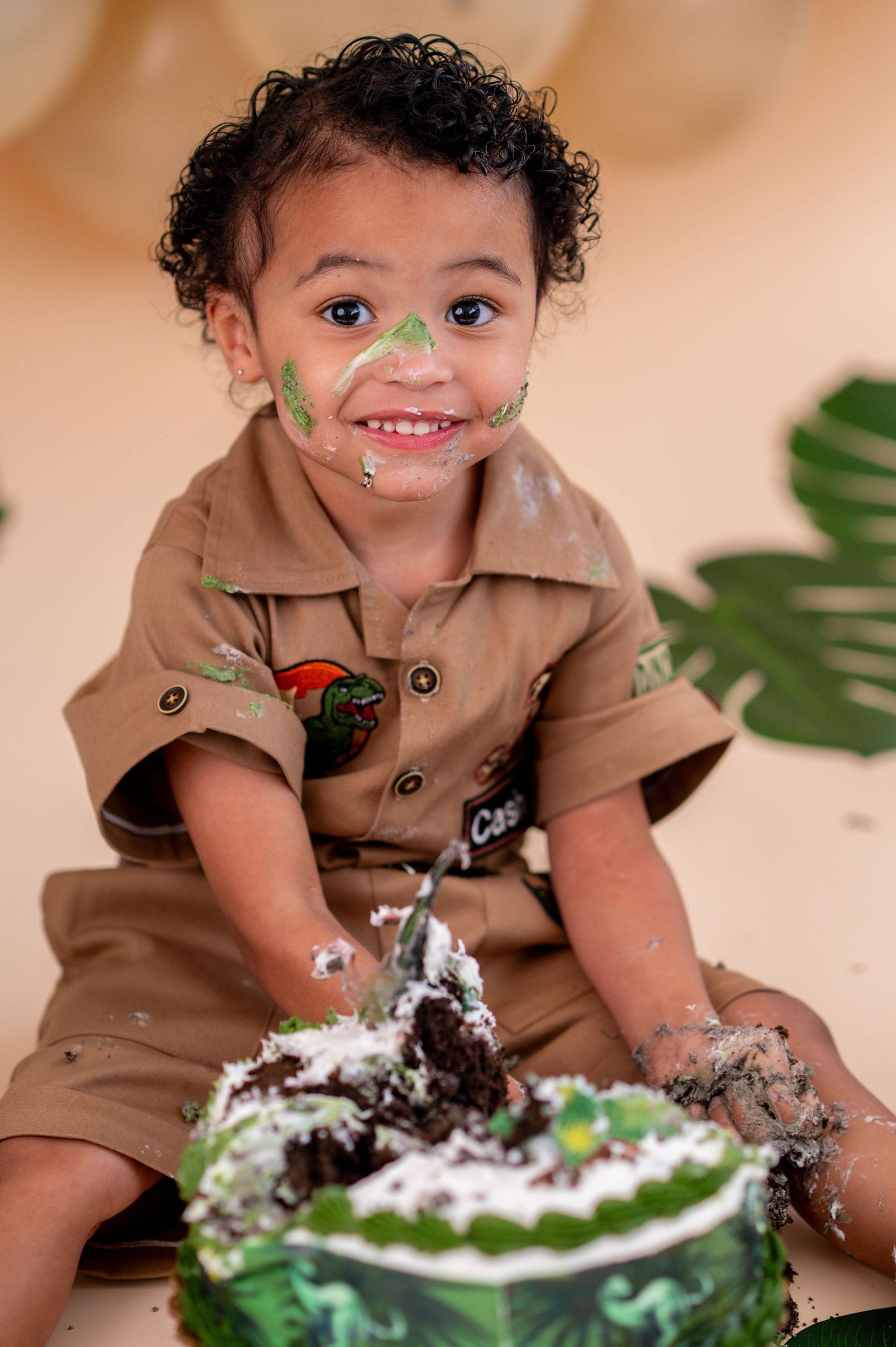 Smiling toddler in khaki outfit with cake smash, green frosting on face, dinosaur-themed birthday cake in front.