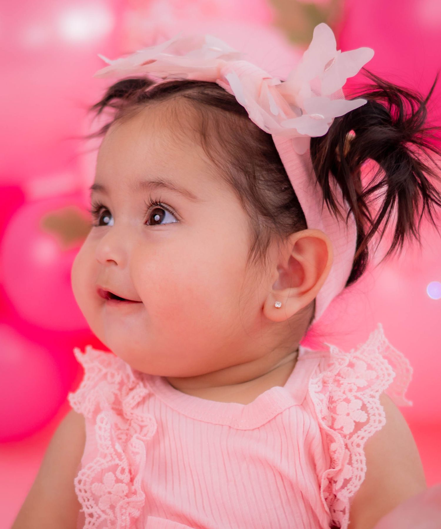 Adorable baby girl in pink outfit and floral headband with pearl earring, smiling against pink balloon backdrop.