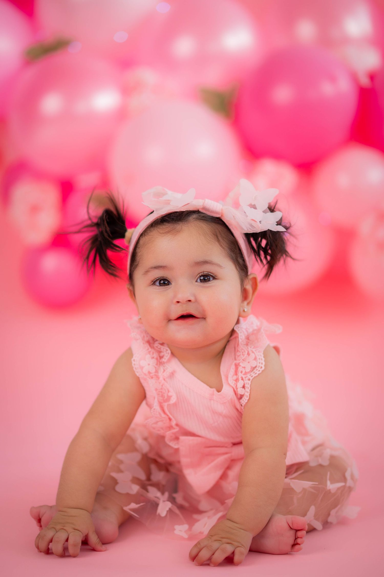 A cute baby girl in a pink ruffled dress and floral headband sits surrounded by pink balloons.