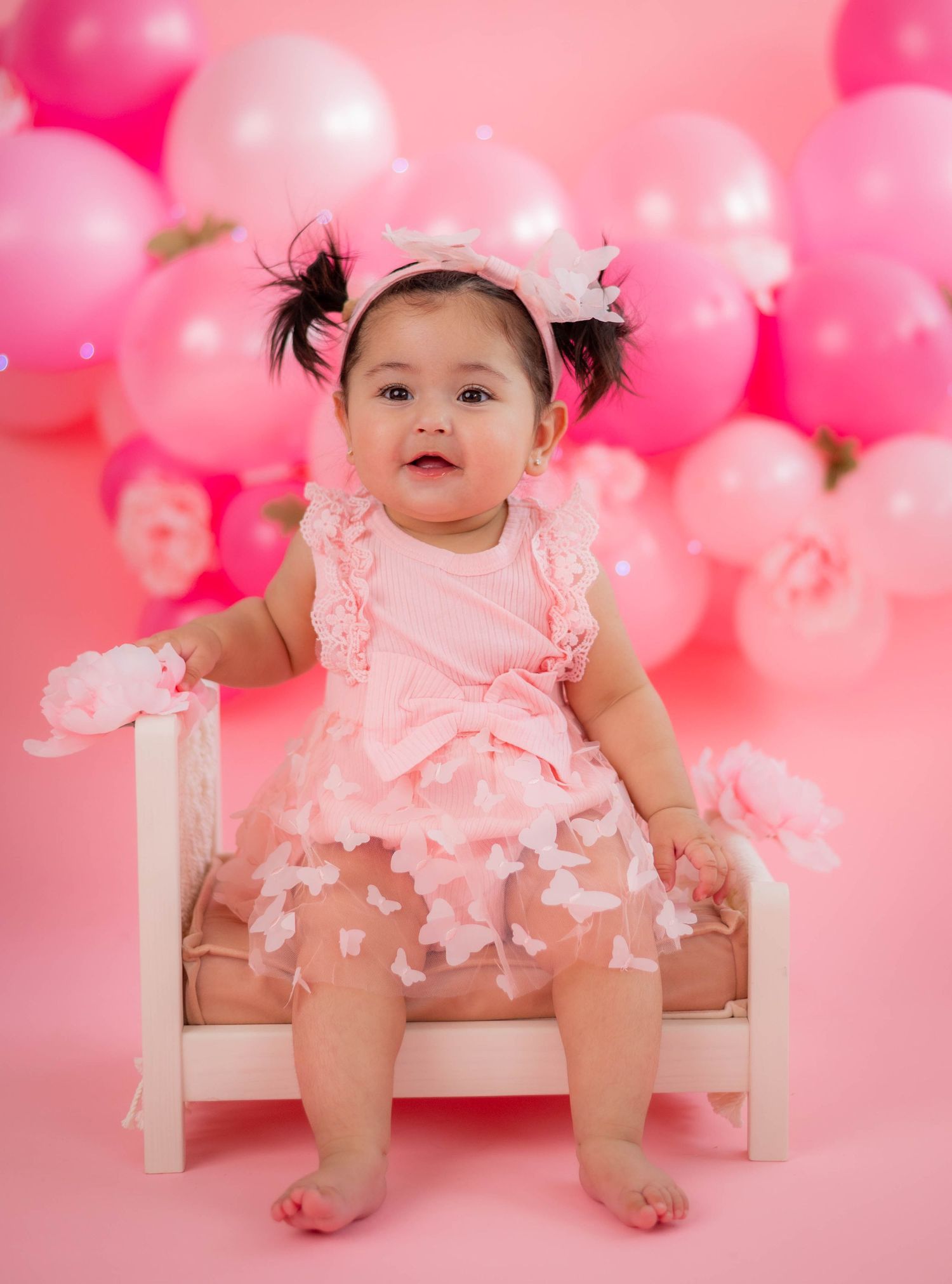 Baby girl in pink dress and bow headband sits on white chair surrounded by pink balloons on pink background.