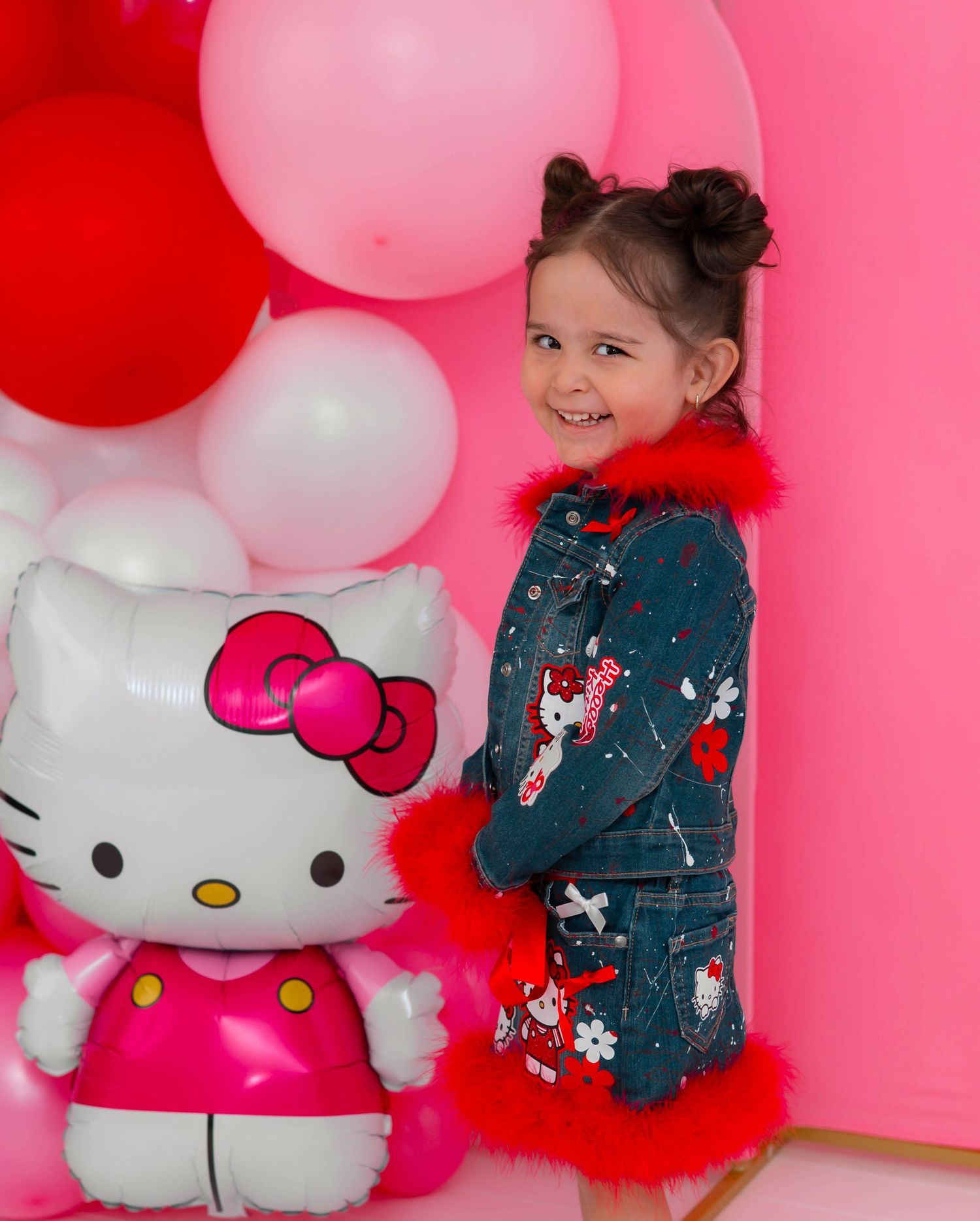 Young girl in Hello Kitty denim jacket with red fur trim smiles beside Hello Kitty balloon at pink party.