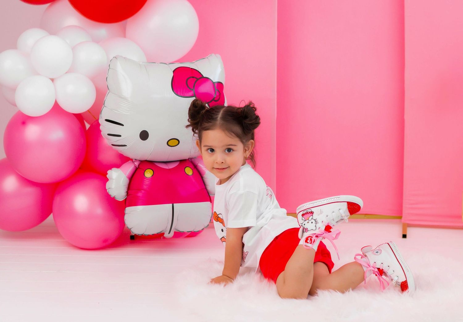 Young girl poses beside a Hello Kitty balloon with pink balloon arch backdrop at a birthday party.