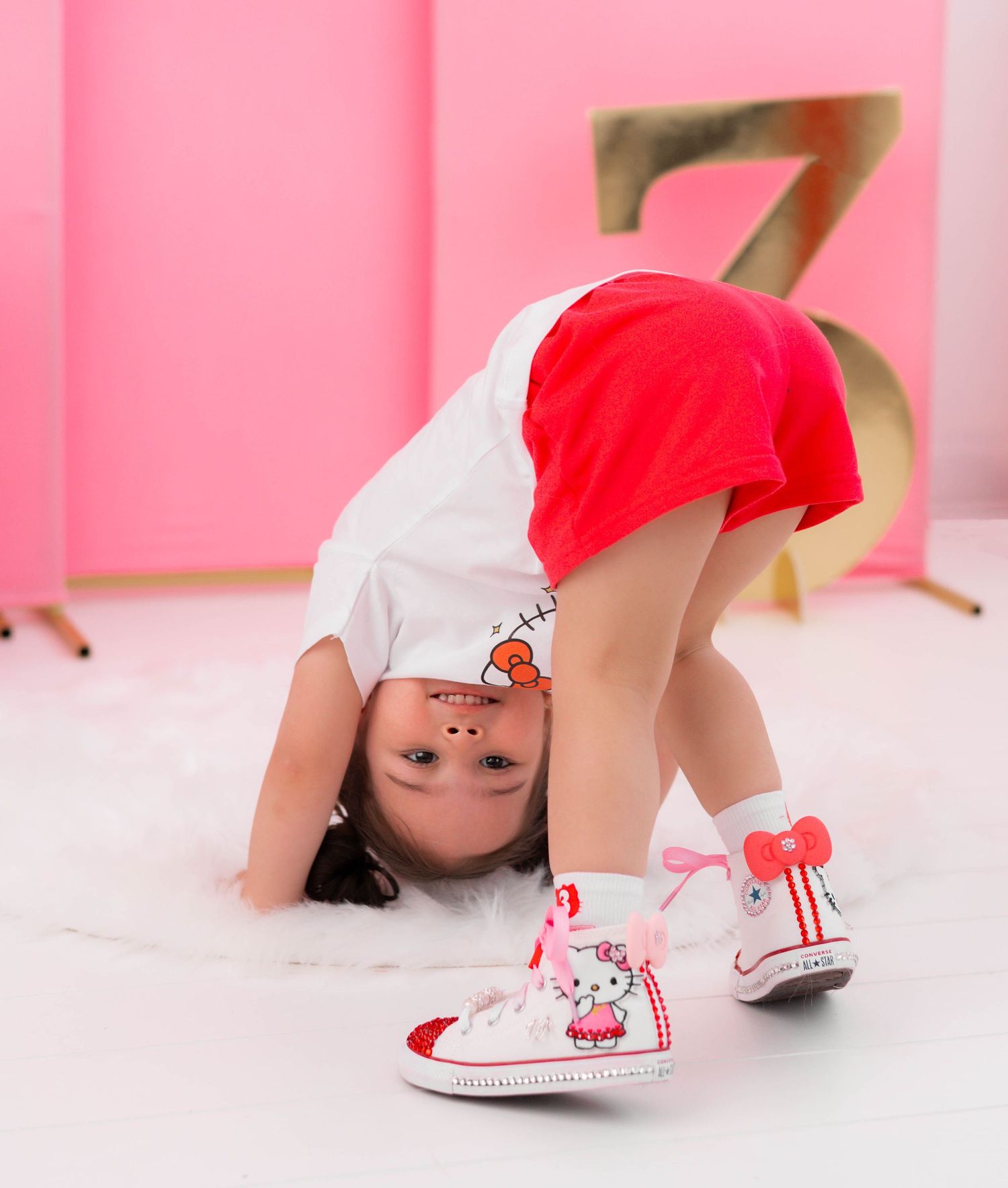 Toddler girl bending over in Hello Kitty sneakers, red skirt, and white tee in a pink studio setting.
