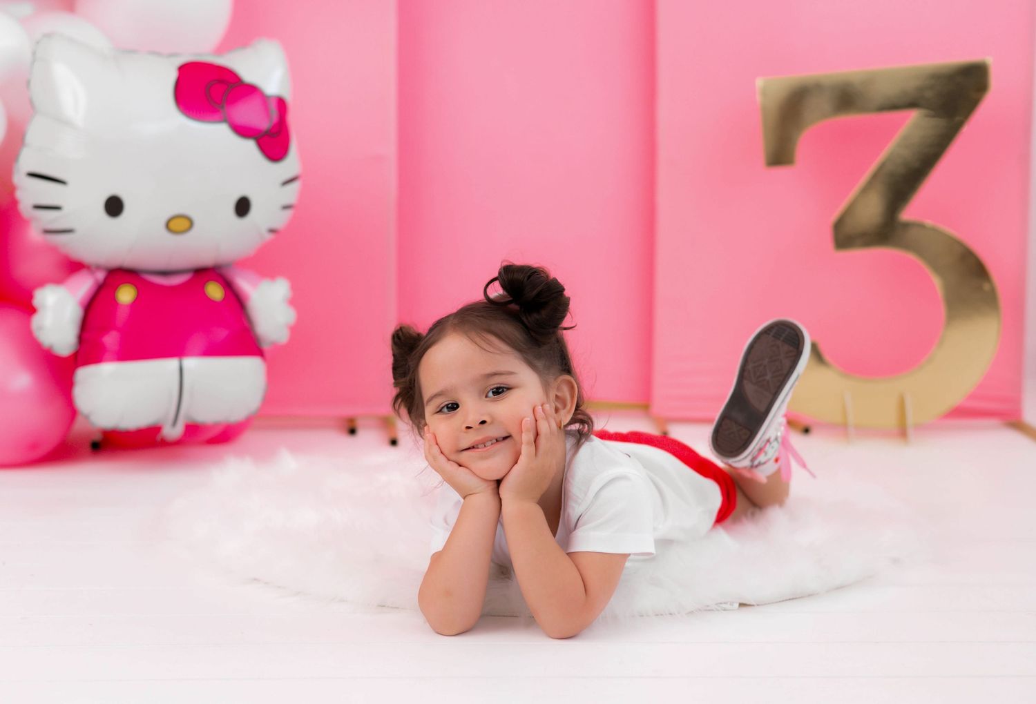Toddler girl poses at Hello Kitty themed 3rd birthday party with pink balloons and gold number three.