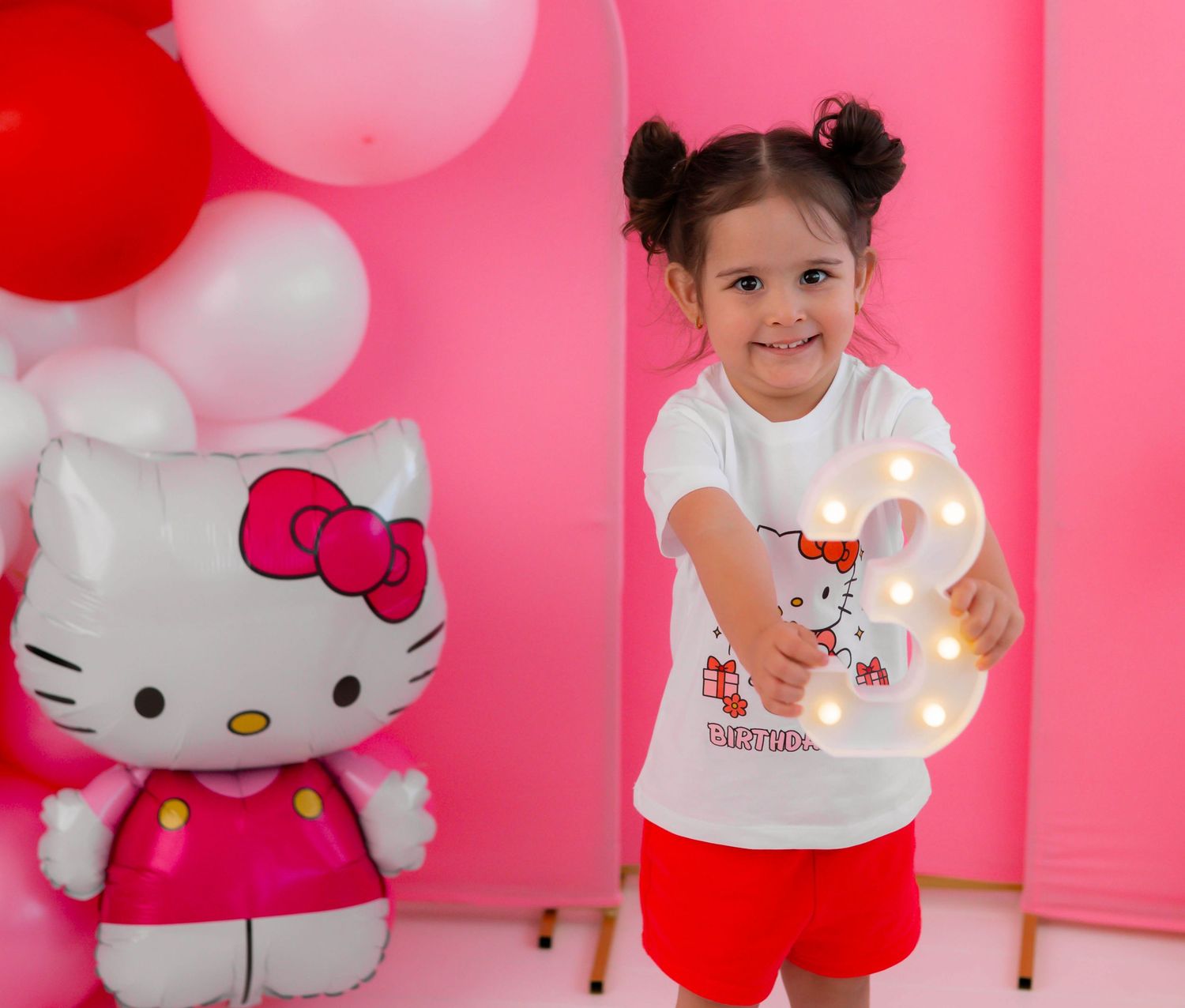 A little girl in a Hello Kitty outfit holds a light-up number 3 at a pink Hello Kitty birthday party.